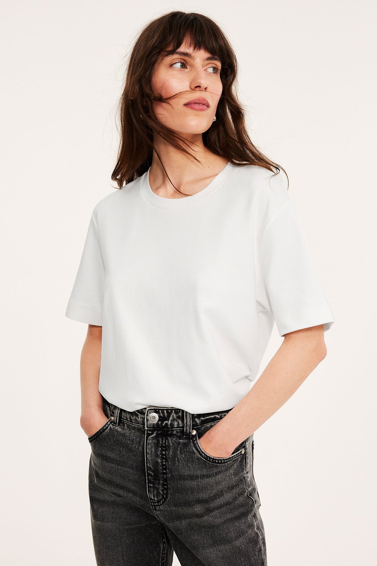 Frontvisning av en kvinne i en offwhite oversized bomulls-t-skjorte og mørkegrå vaskede jeans.