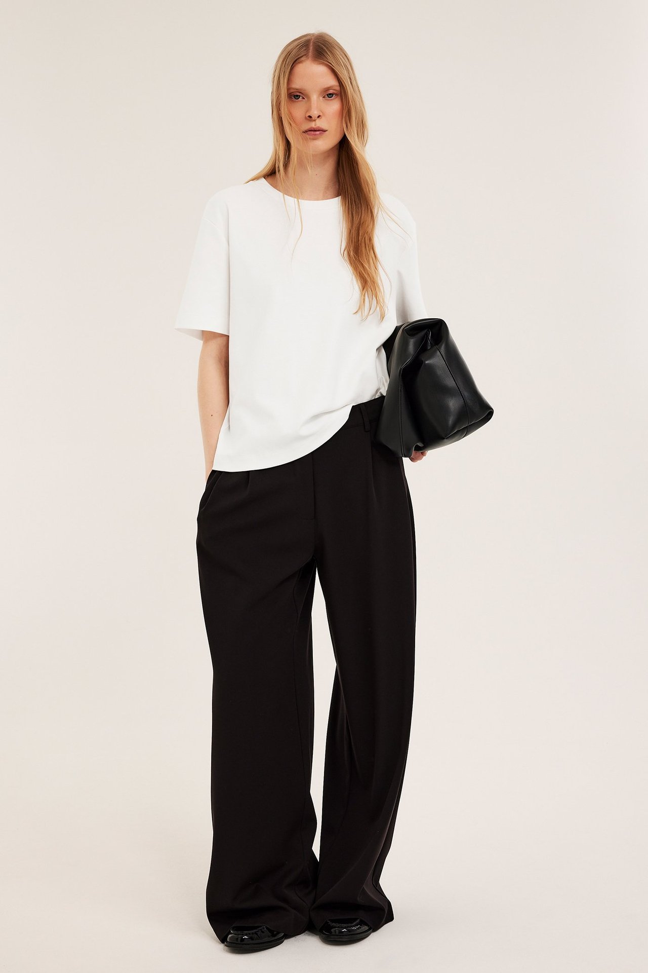 Framifrån: Kvinna i oversized offwhite t-shirt i bomullsinterlock, svarta vida byxor, svarta platta skor, med svart handväska.