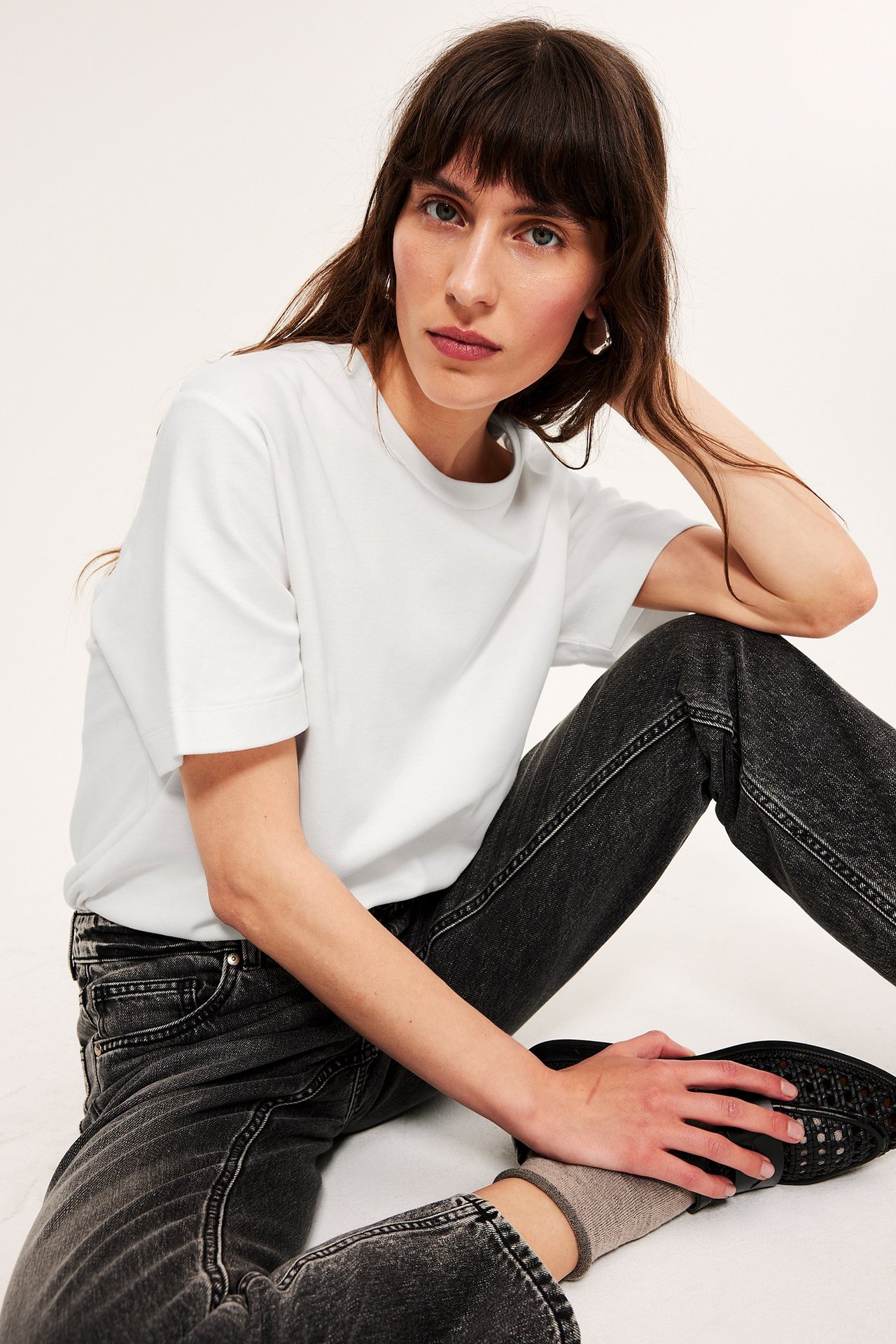 Forfra: kvinne i offwhite oversized bomulls-t-skjorte, svarte jeans, sølvøreringer og svarte sko.