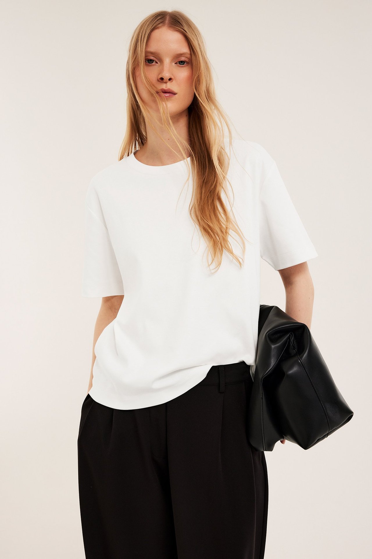 Widok z przodu kobiety w obszernej bawełnianej koszulce ecru, czarnych spodniach i z czarną torebką.