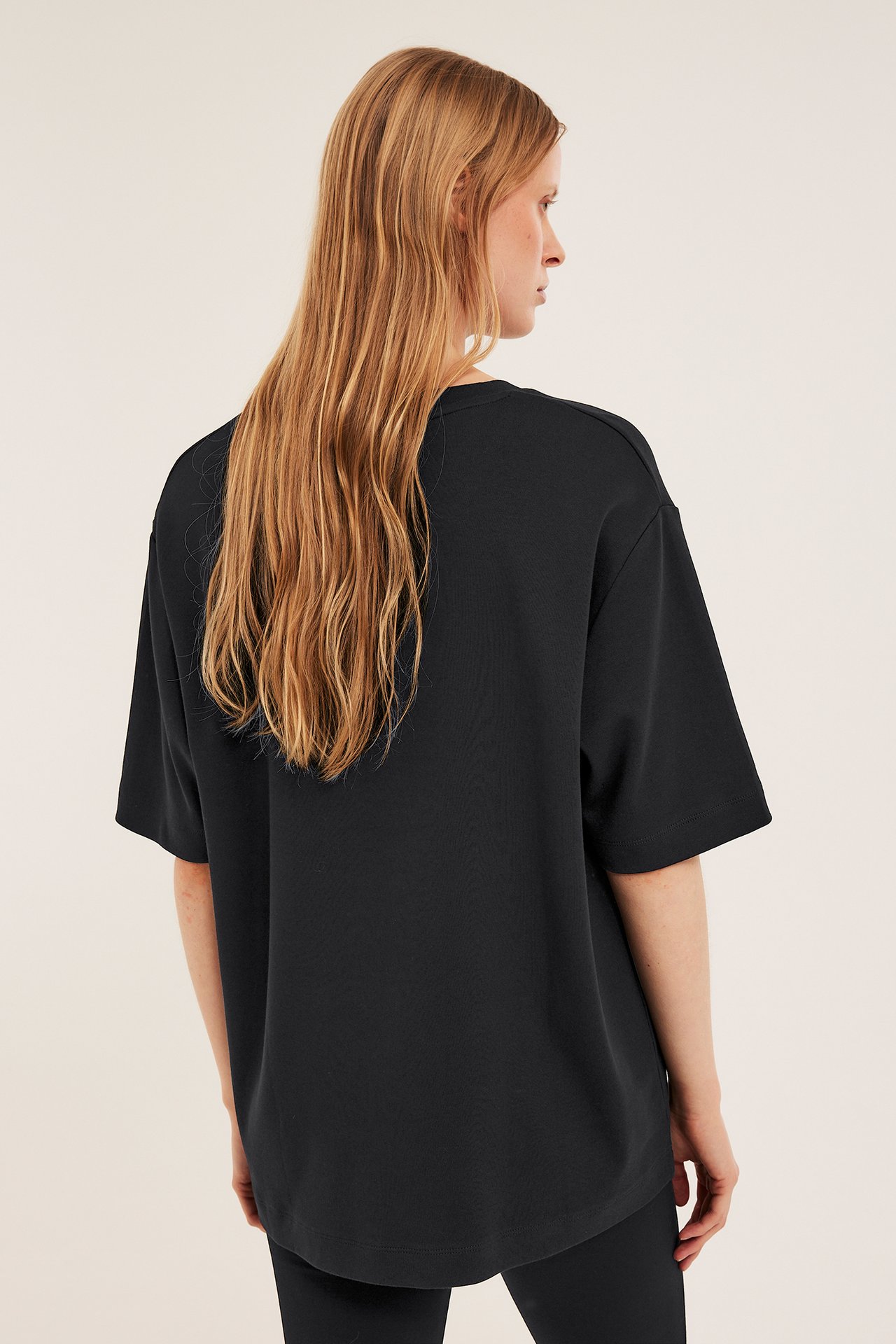 Ryggvy av kvinna i svart, enfärgad oversized T-shirt i bomullsinterlock och svarta byxor.