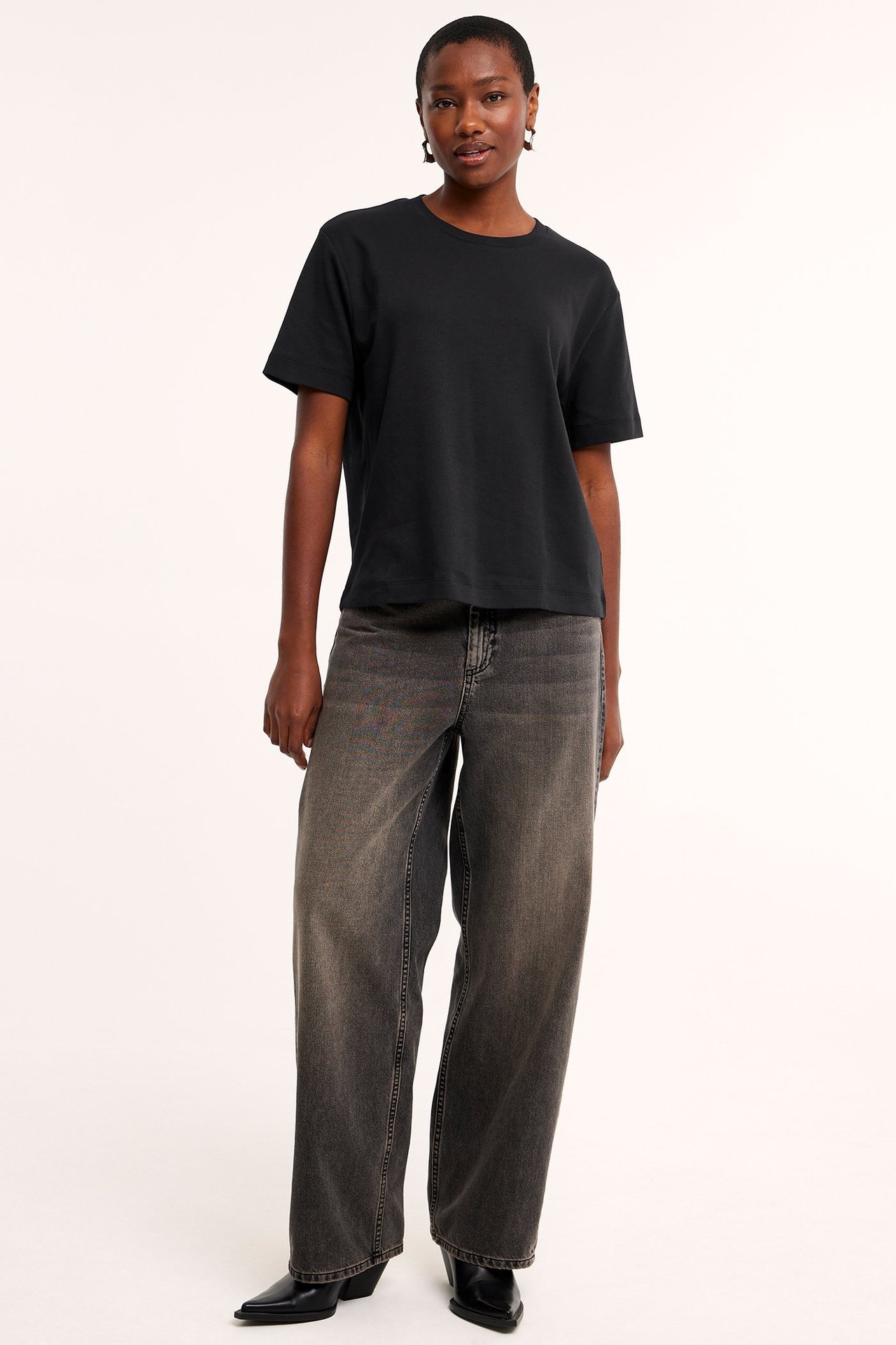 Forfra: kvinne i svart oversized bomulls-T-skjorte, mørkegrå vide jeans og svarte spisse støvler.