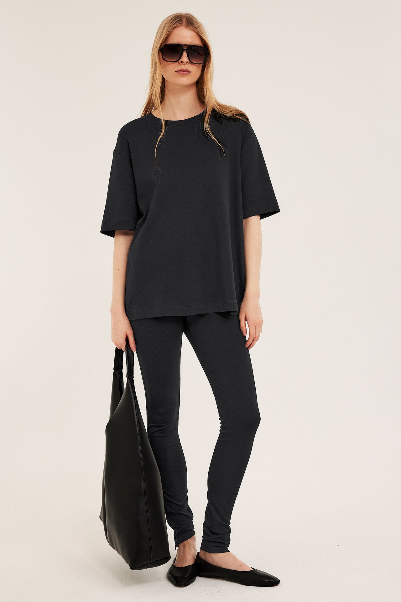 Frontvy: Kvinna bär en svart oversized interlock t-shirt, svarta leggings, svarta solglasögon, håller en svart väska.