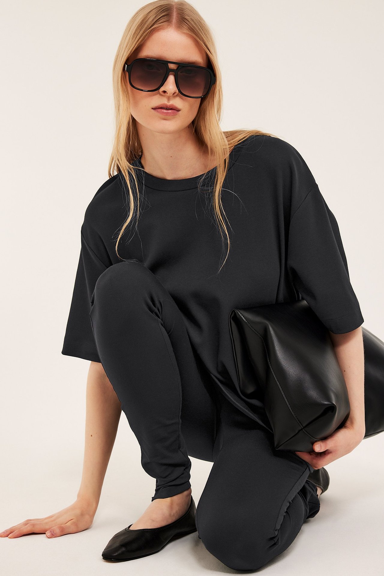 Kvinne i oversized svart t-skjorte, svarte leggings, ballerinasko, solbriller, holder en svart veske.