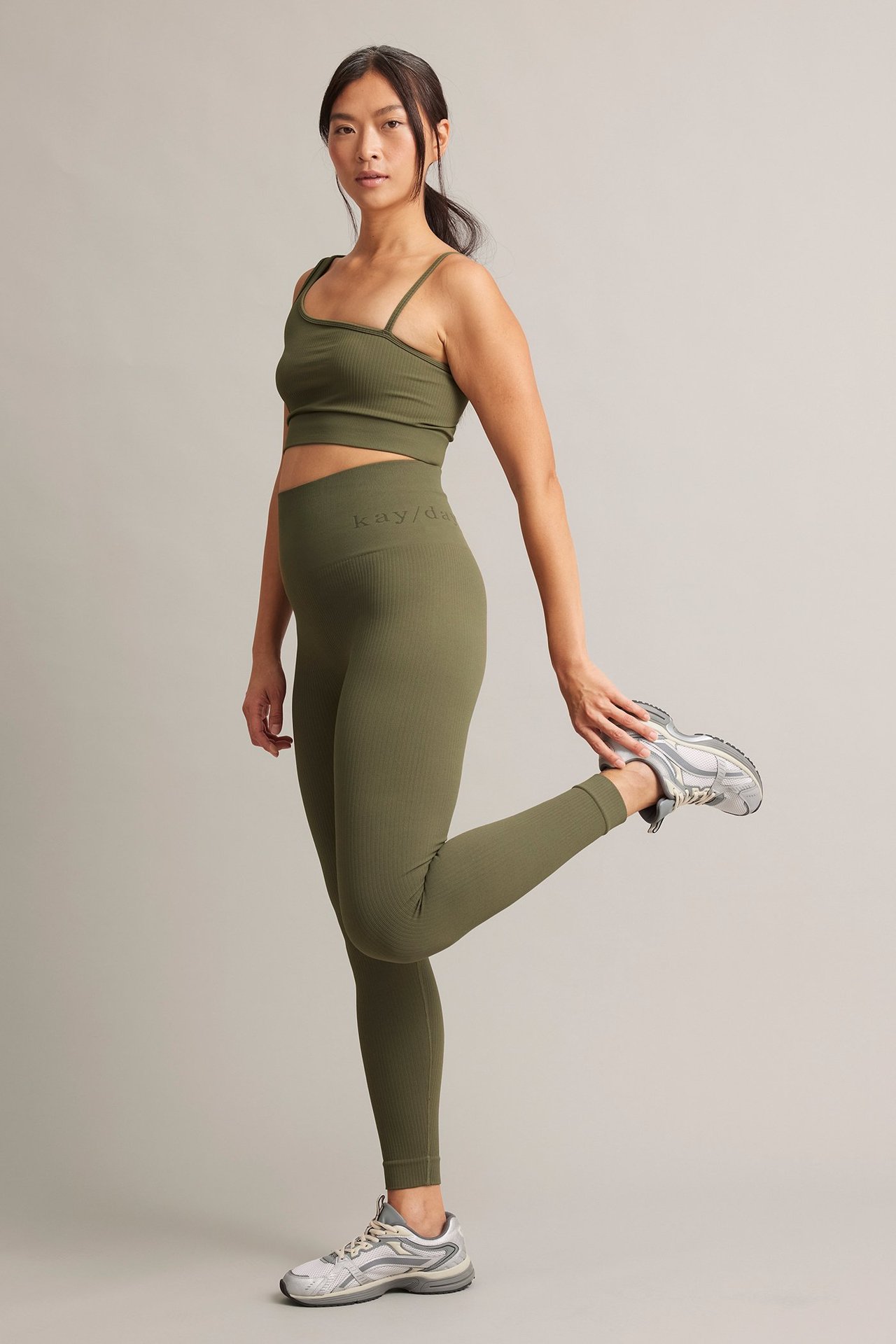 Framifrån, kvinna i olivgröna ribbade träningstights, matchande sport-bh och silverfärgade träningsskor.