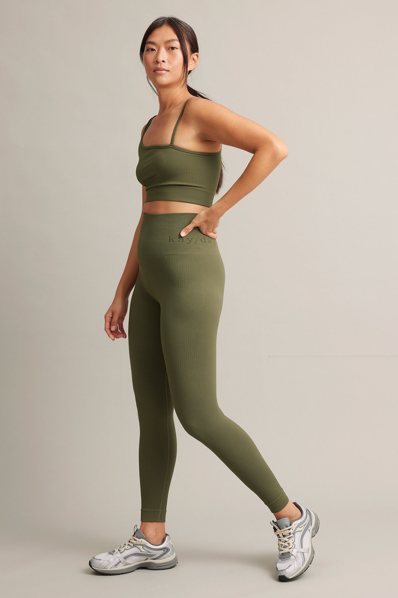 Sidebilde av en kvinne i grønn ribbet asymmetrisk sportstopp, matchende leggings og grå joggesko.