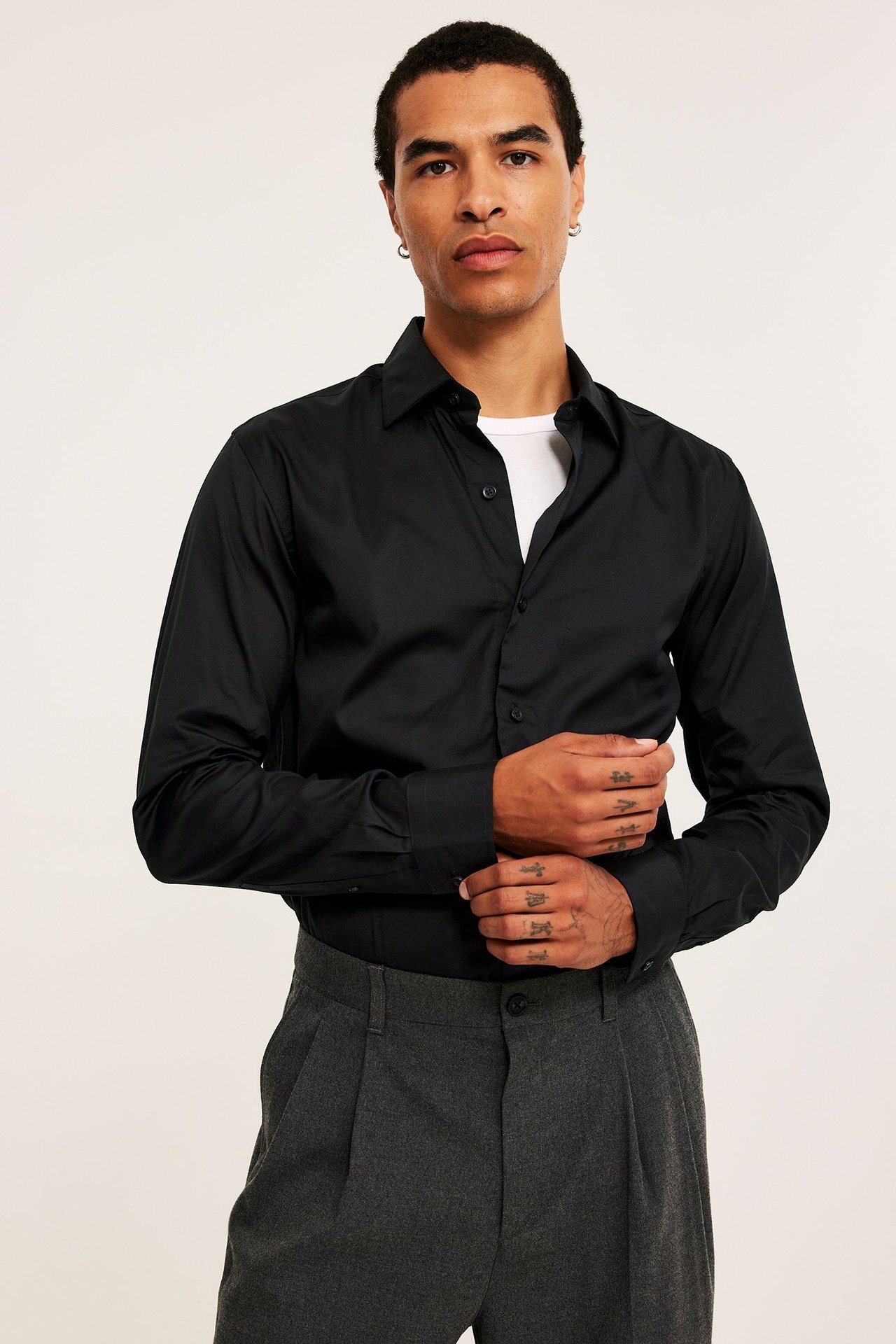 Frontvy av en man i svart slim fit bomullsskjorta över vit t-shirt och grå plisserade byxor.