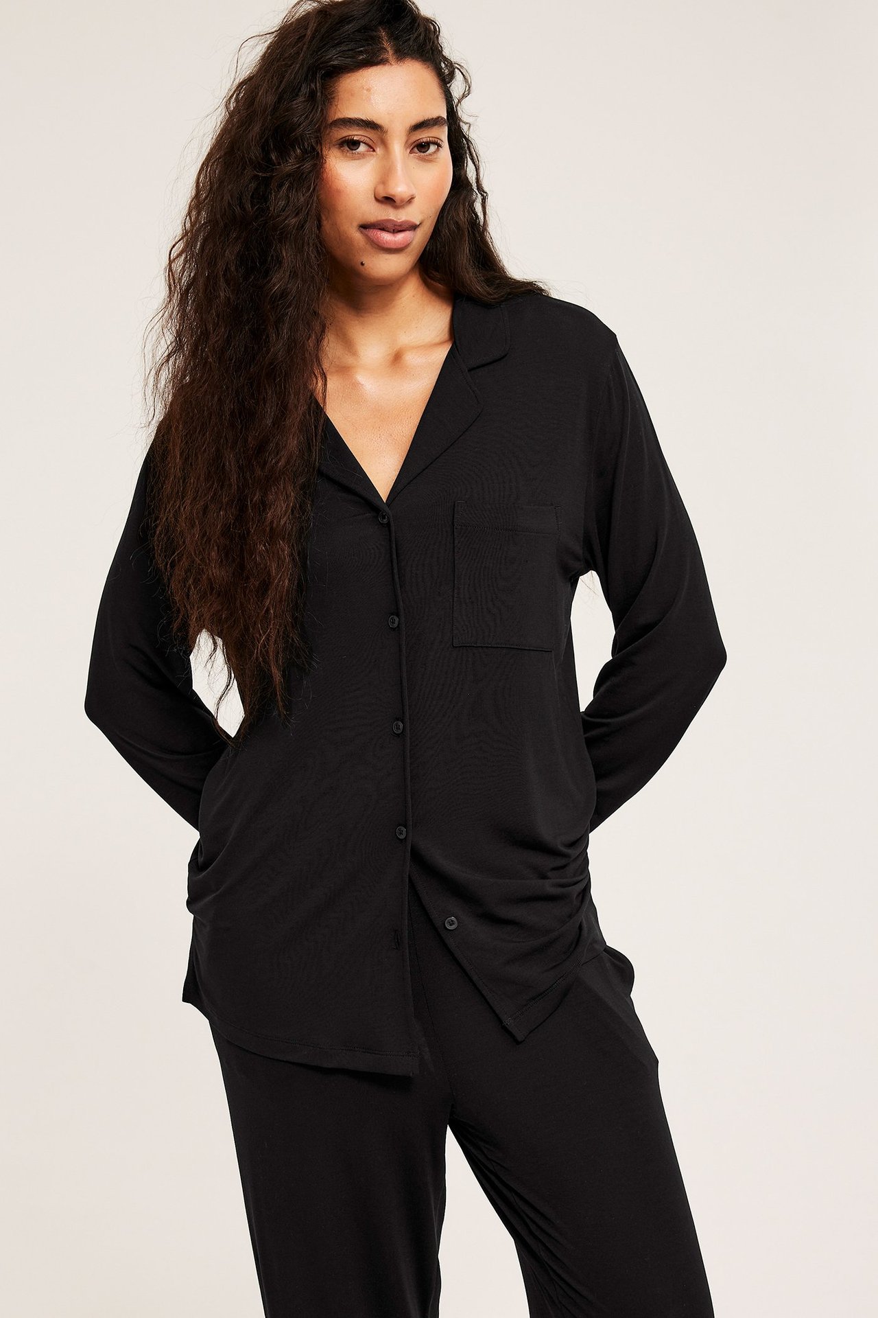 Forfra: en kvinne i svart lyocell-pyjamas med langermet knappeskjorte og avslappede bukser.