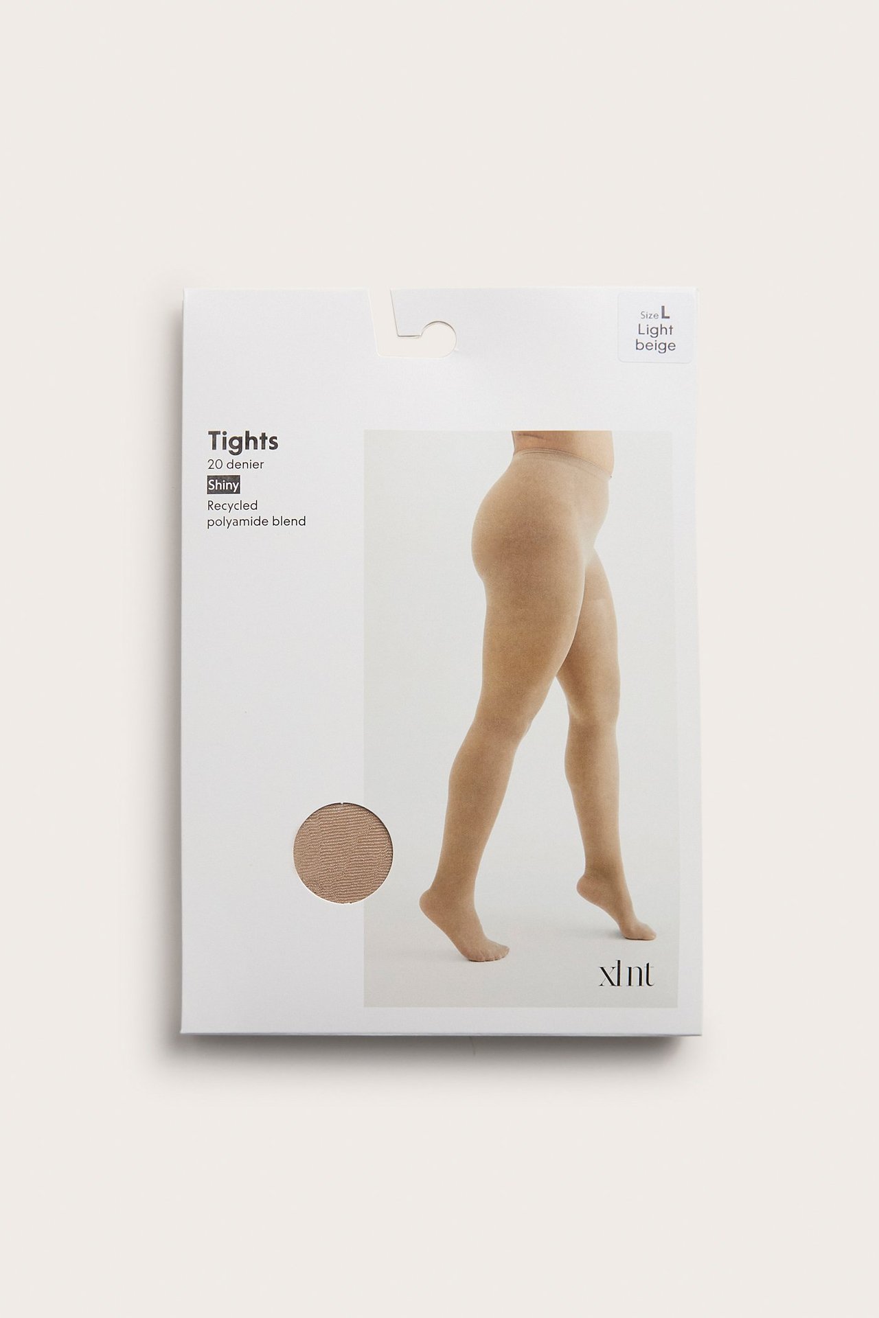 Edestä näkymä valkoisesta pakkauksesta vaaleanbeigelle kiiltävälle 20 denierin sukkahousuille, naisen jalassa.