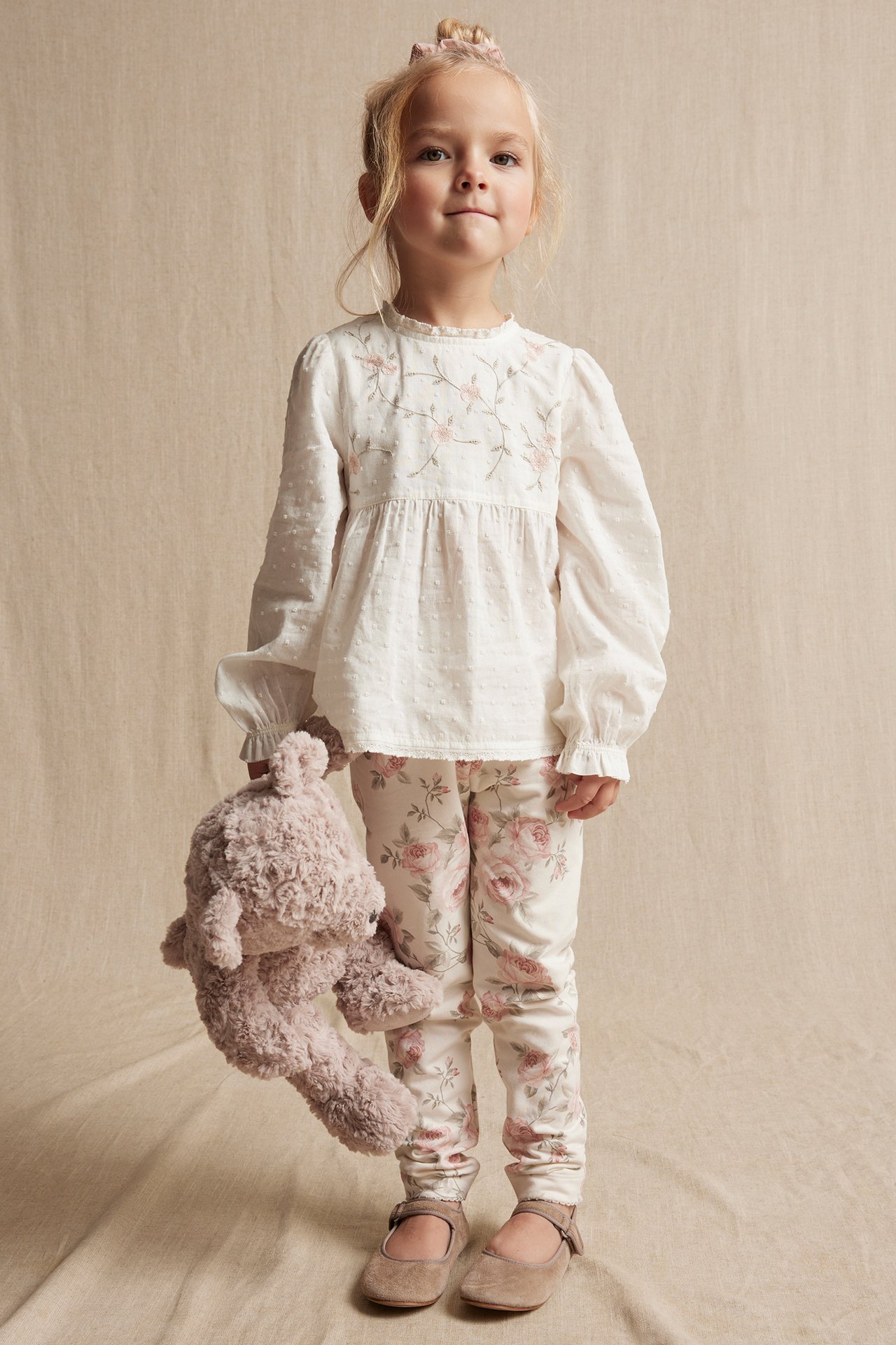 Barn i offwhite brodert bomullsbluse, blomstrete leggings, brune sko, holder kosedyr.