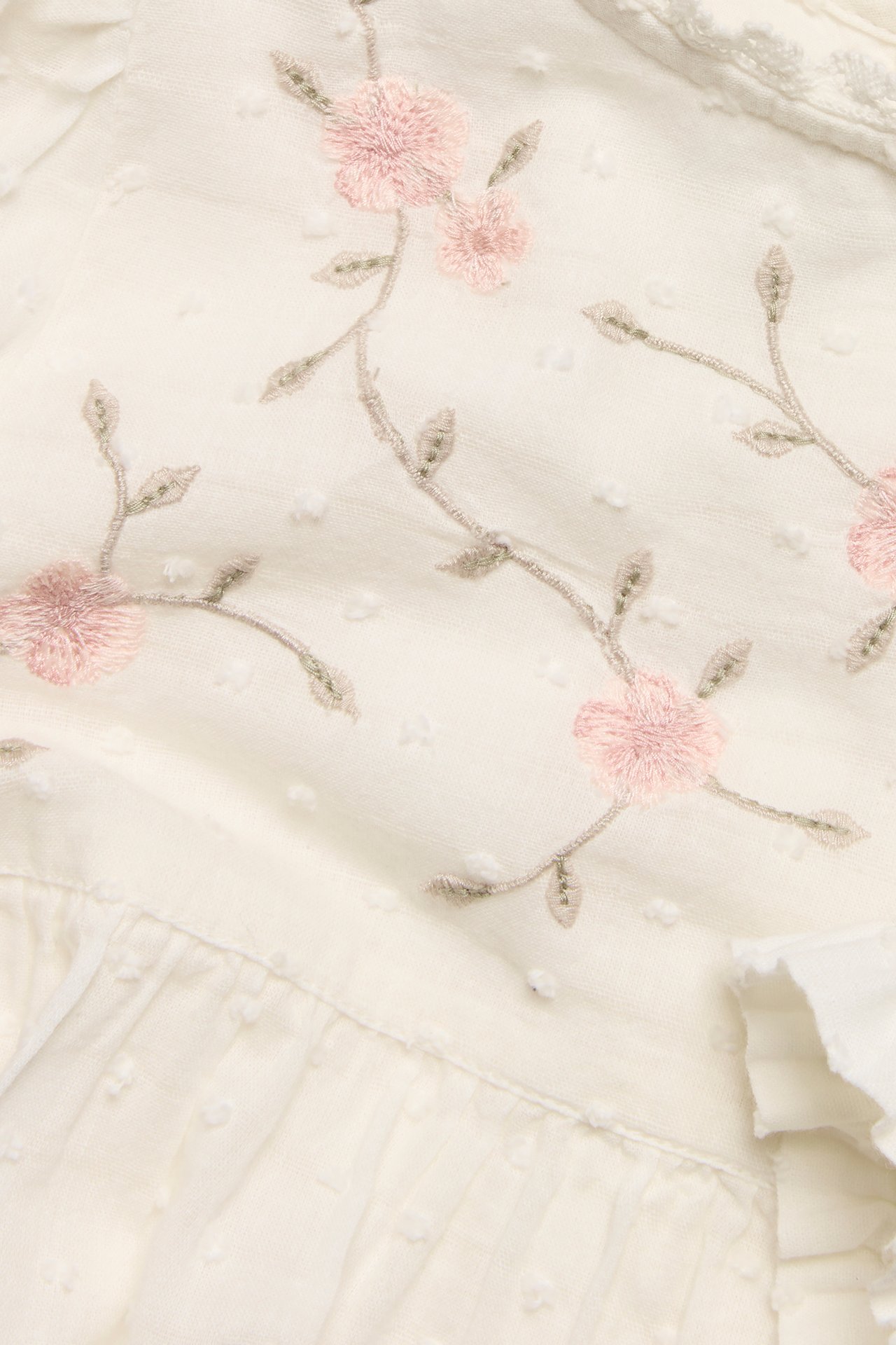 Nærbilde av en offwhite vevd bomullsbluse med rosa blomsterbroderi, teksturerte prikker og volangkant.