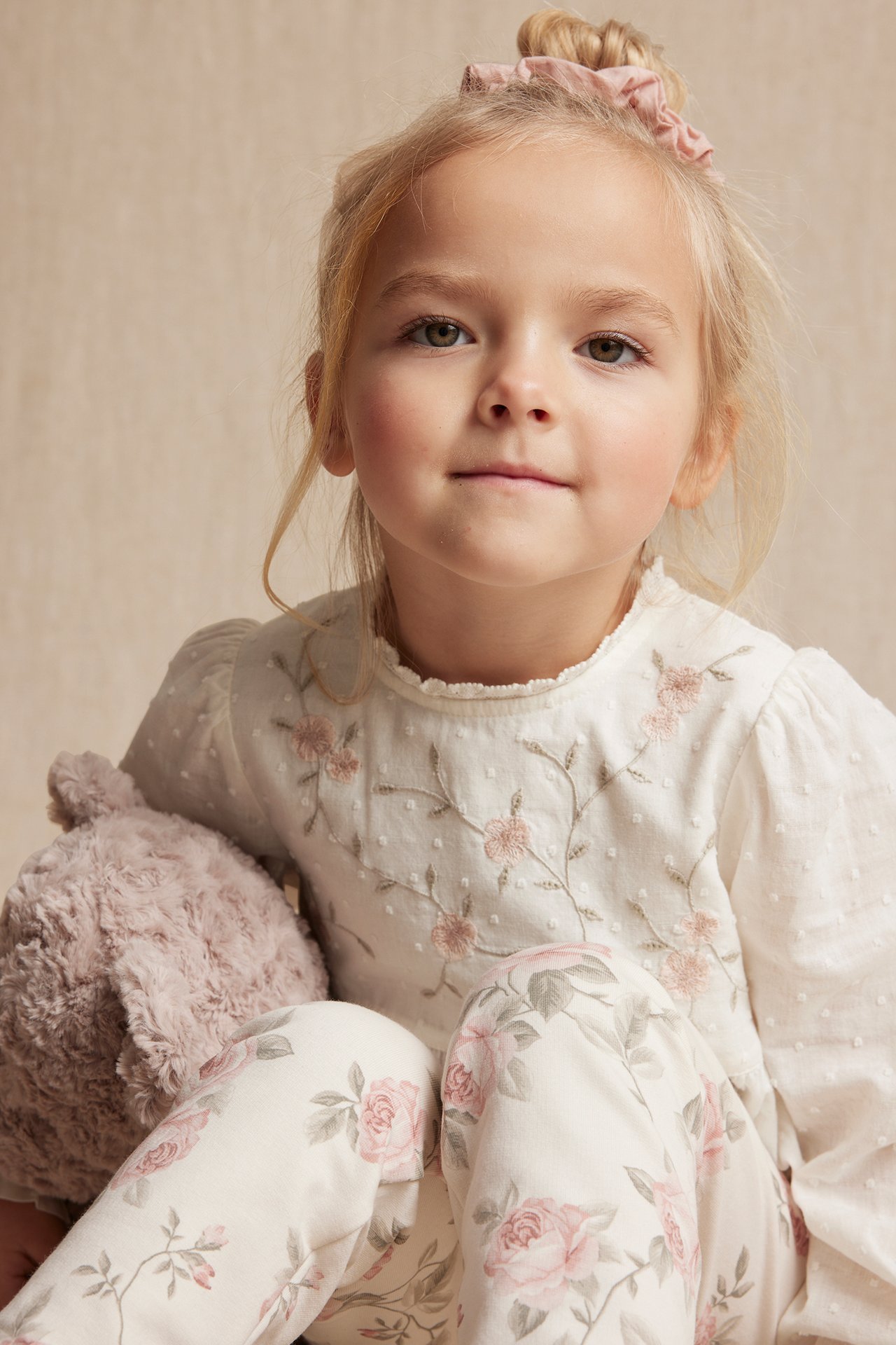 Nærbilde av et barn i en offwhite brodert bomullsbluse, blomstrete bukser og en rosa scrunchie.