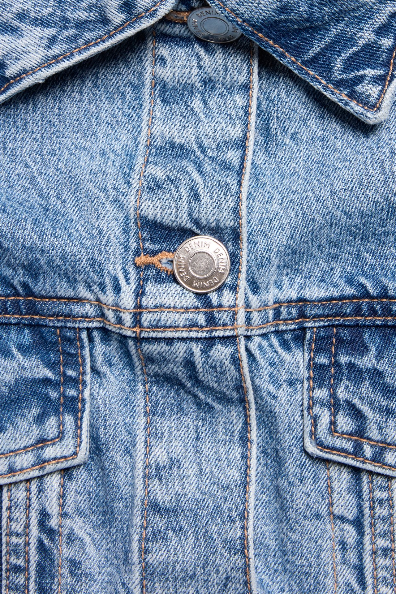 Närbild av en blå tvättad oversized jeansjacka med kontrastsömmar, metallknappar, krage och bröstfickor.