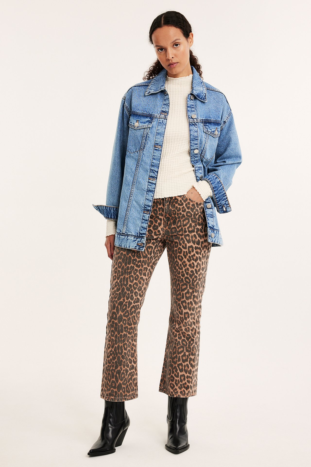 Framifrån: Kvinna i oversized ljusblå jeansjacka, krämvit ribbad topp, leopardmönstrade croppade jeans, svarta stövlar.