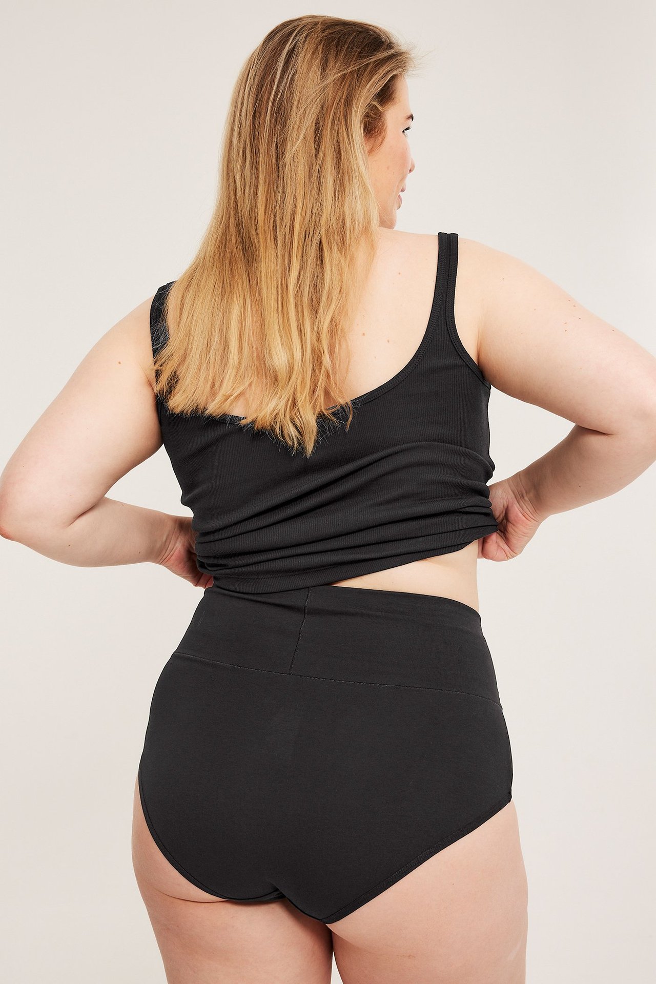 Bakifrån, kvinna i svarta bomullshipstertrosor med brett midjeband och ett svart ribbat linne.