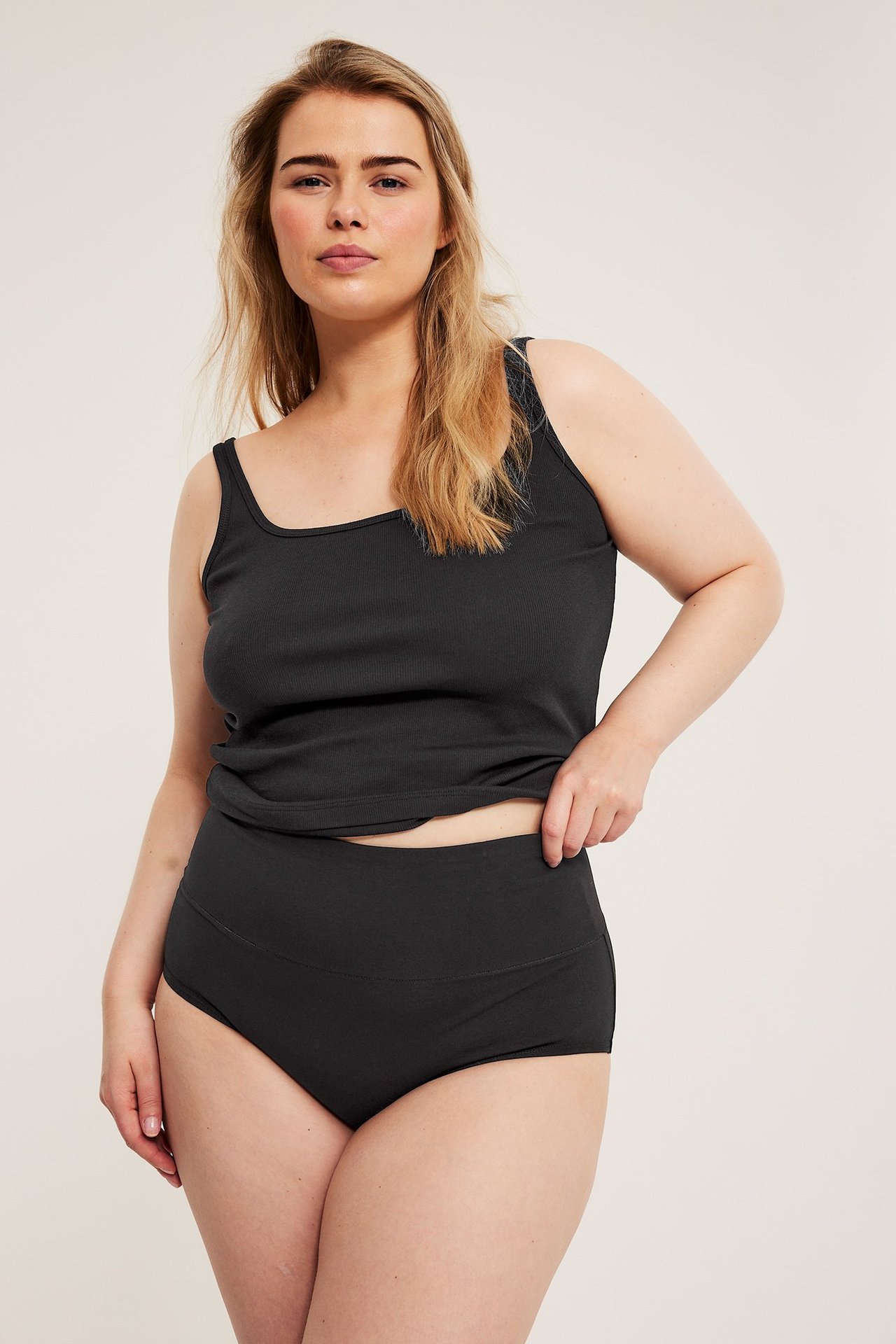 Framifrån, kvinna i svarta bomullshipstertrosor med brett midjeband och ett svart ribbat linne.