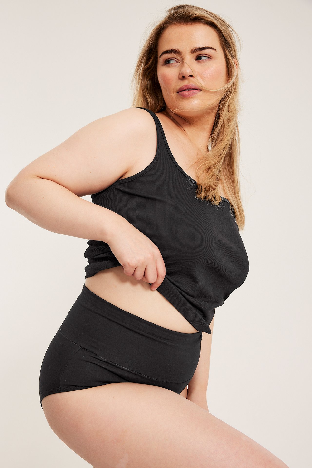 Sidovy av kvinna i svarta bomullshipstertrosor med brett midjeband och svart ribbad tanktopp.