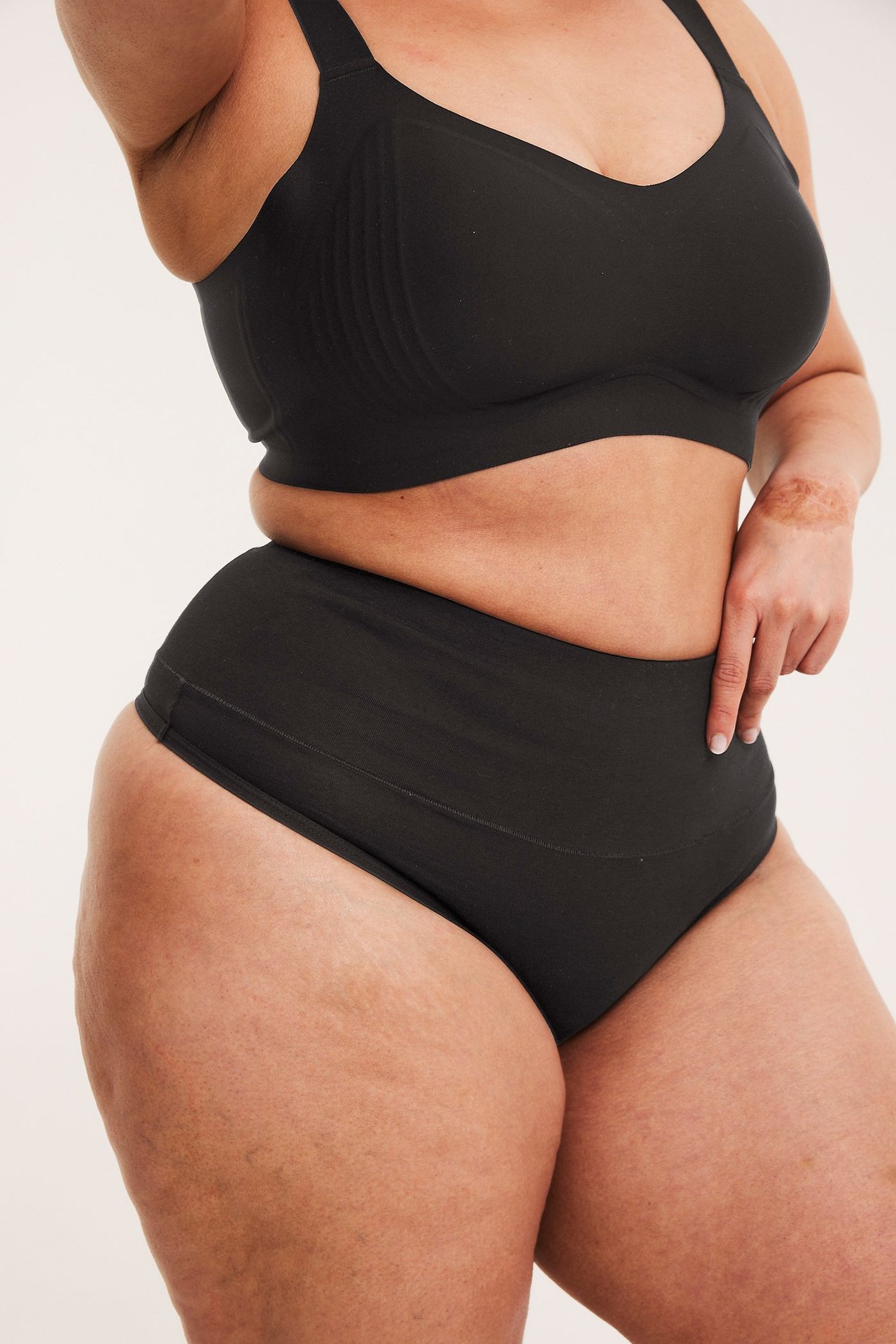 Sidebilde av kvinne i svart bomullstringtruse med bredt midjebånd og svart BH.