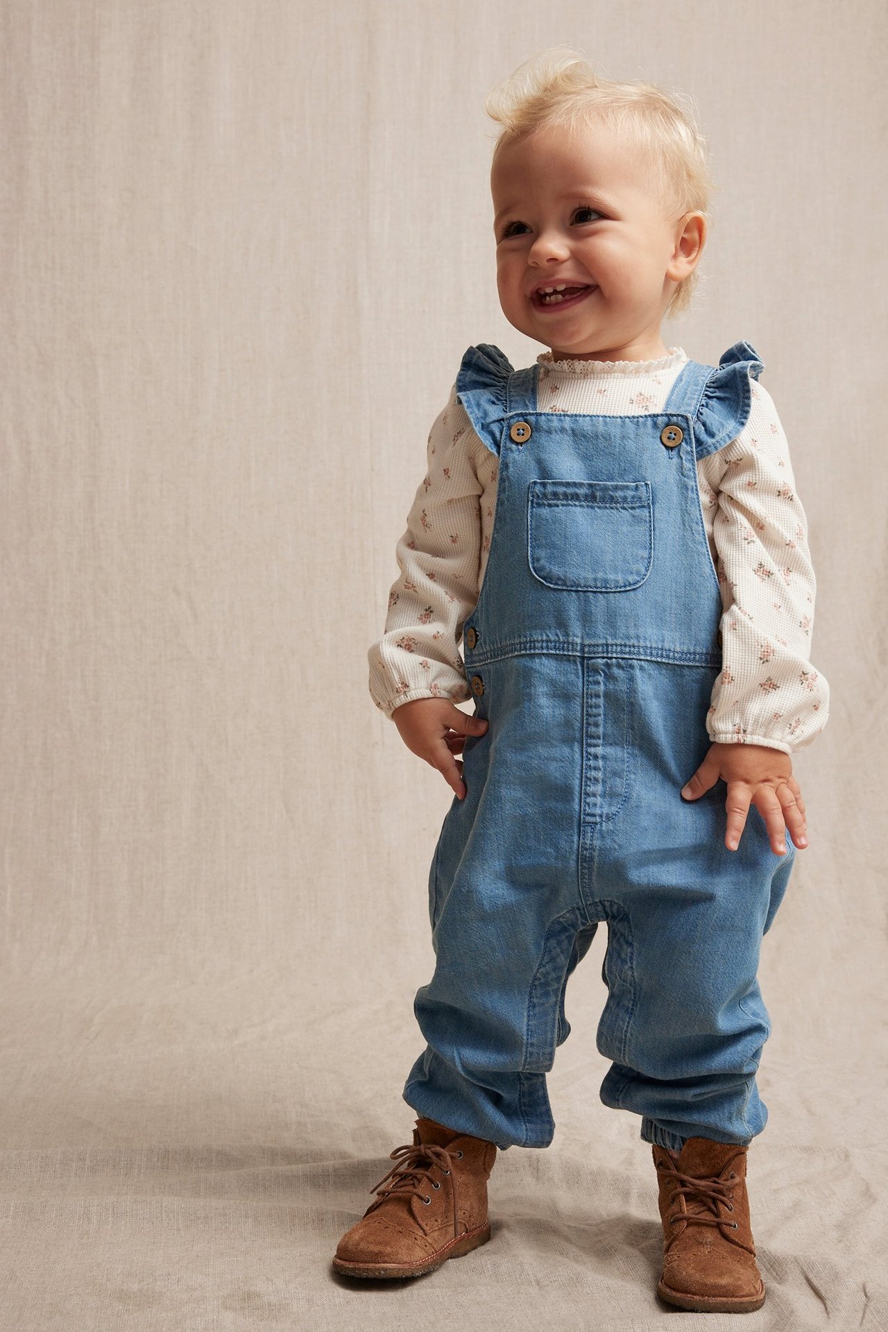 Edestä: Vauva vaaleansinisissä denim-haalarihousuissa, joissa röyhelöolkaimet, kermanvärinen kukkatoppi ja ruskeat nauhakengät.