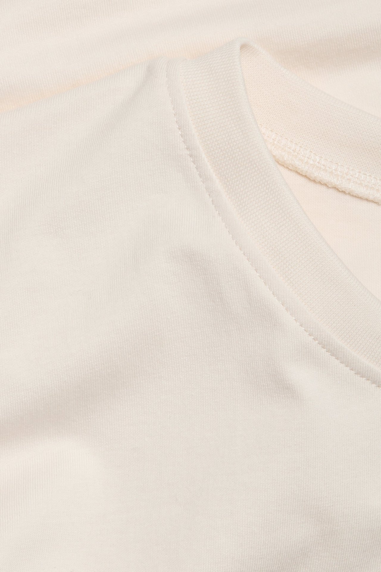 Nærbilde av en offwhite T-skjorte i bomull, som viser den ribbestrikkede rundhalsen og skuldersømmen.