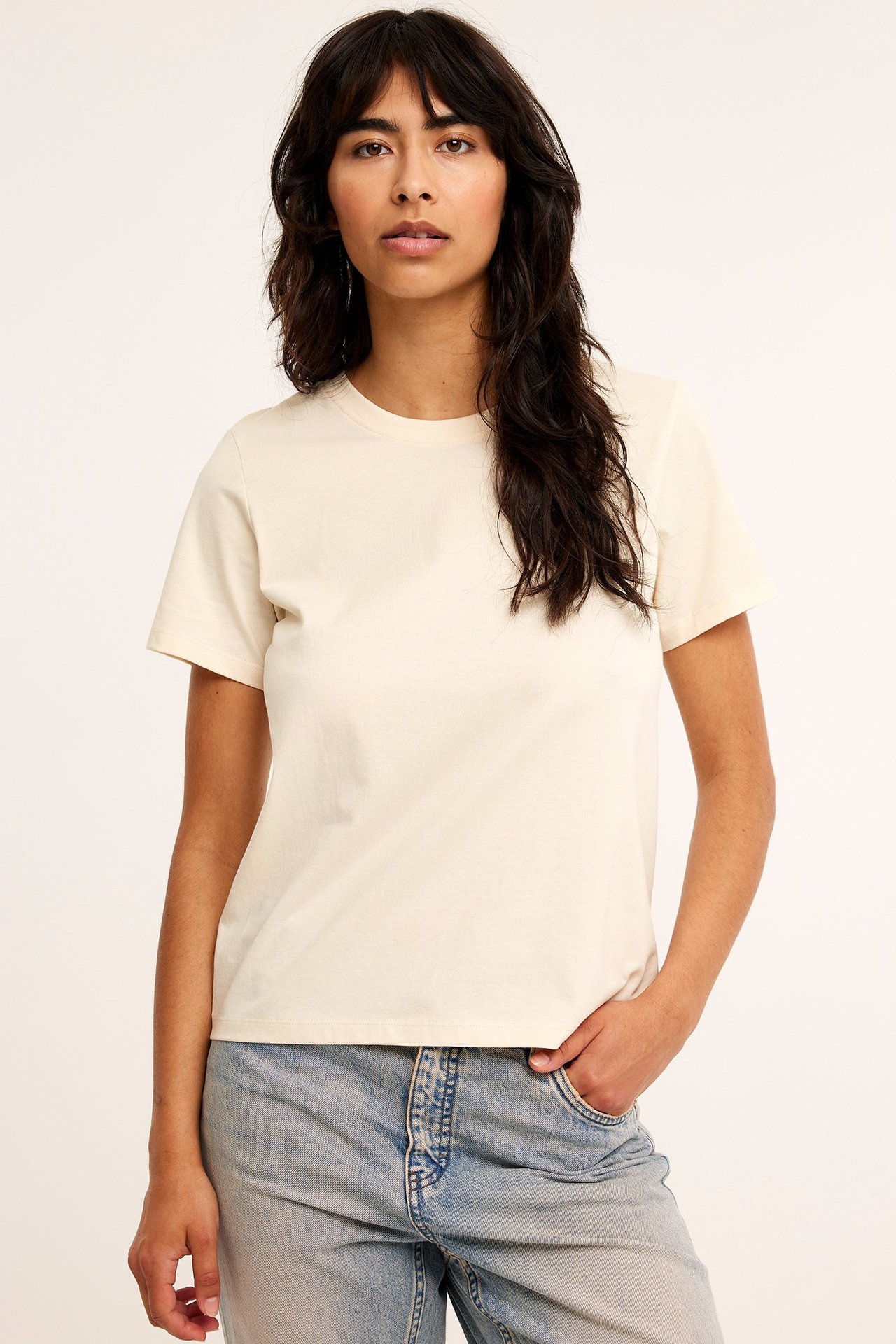 Forfra: kvinne i en kremhvit, ensfarget, rett bomulls-T-skjorte og lyseblå jeans.