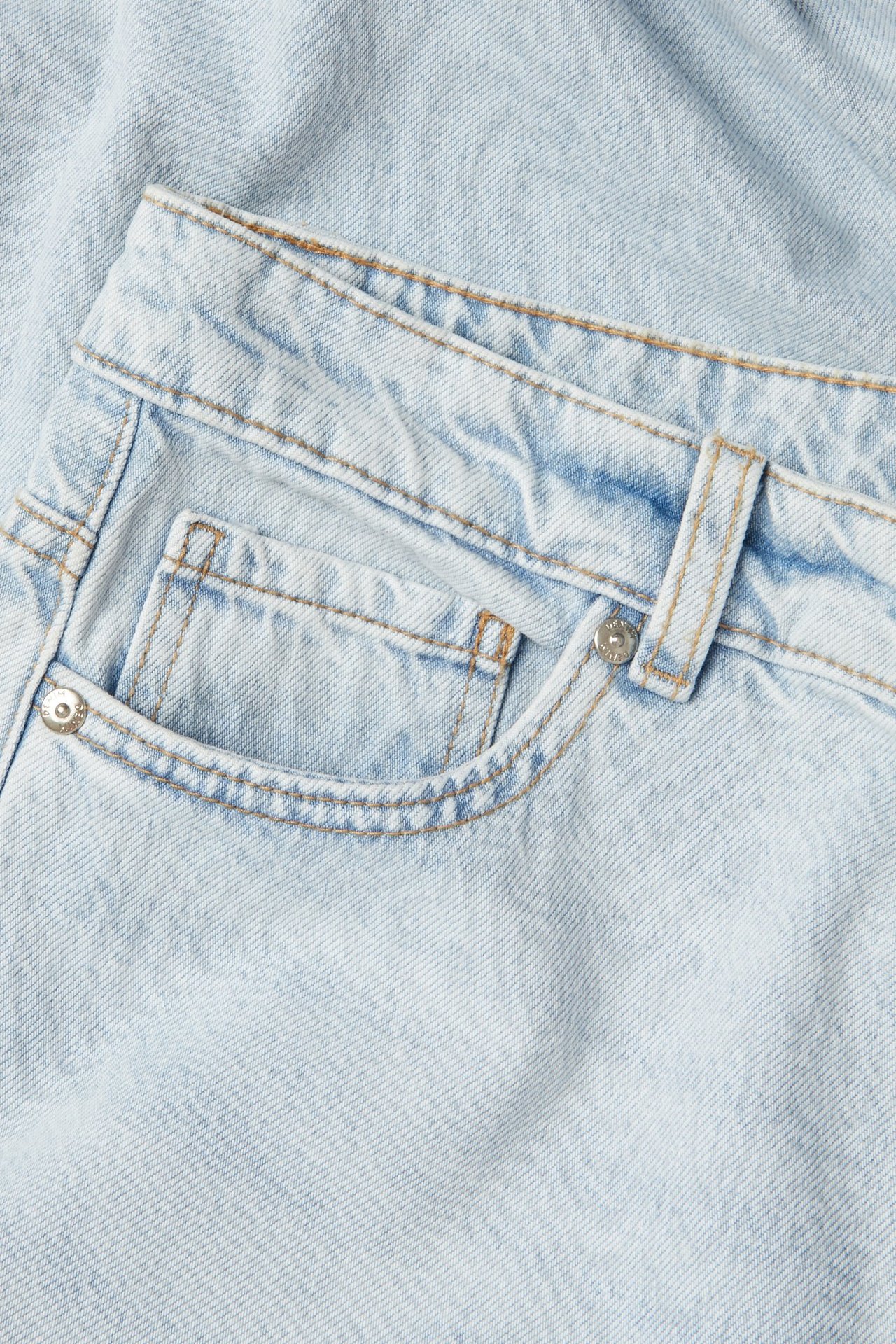Zbliżenie jasnoniebieskich dżinsów z wysokim stanem, szerokimi nogawkami, pokazujące przednią kieszeń, pomarańczowe szwy i nit.