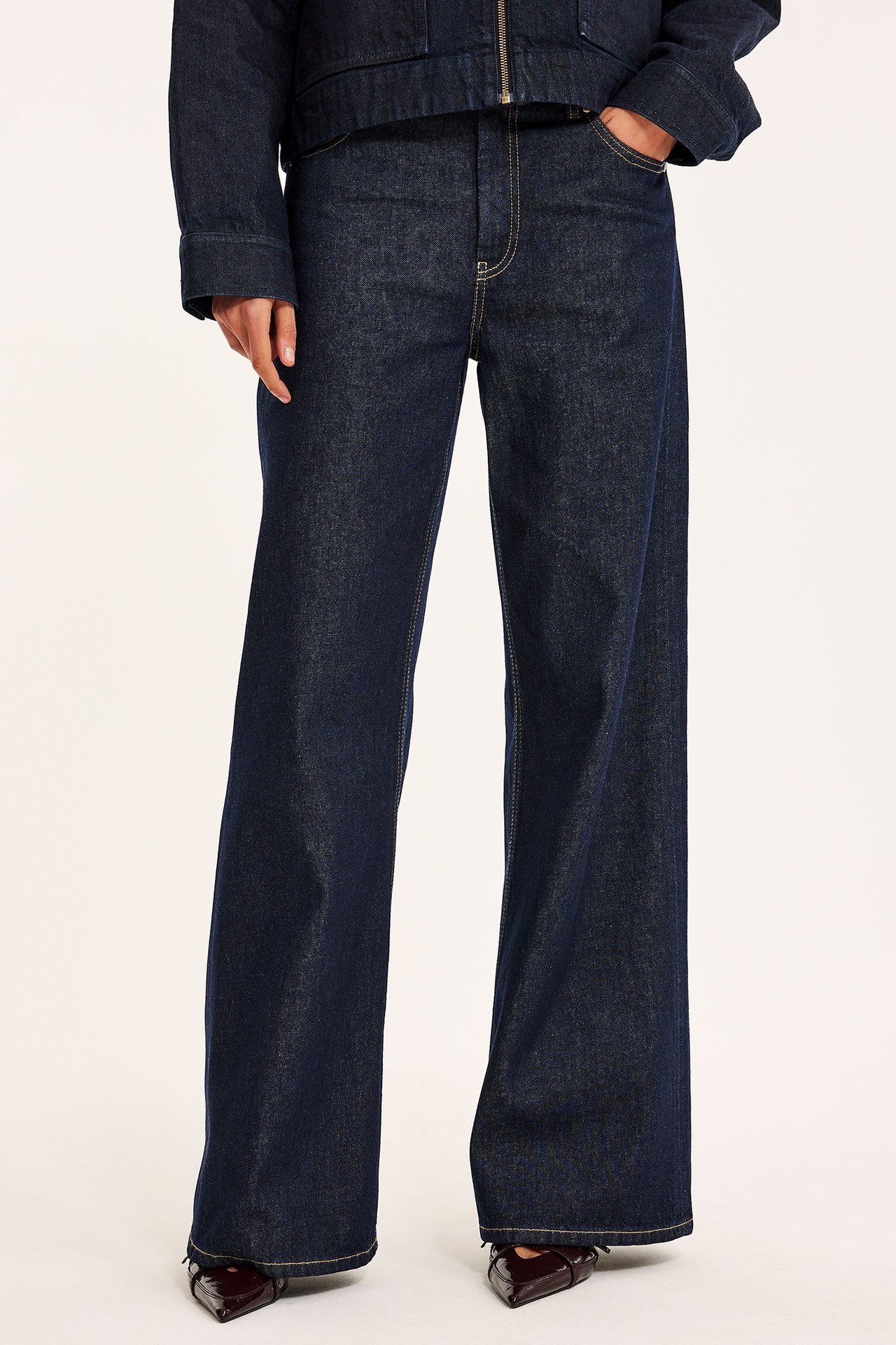 Edestä näkymä naisesta yllään tummat denim high-waist leveälahkeiset farkut, tumma denimtakki ja tummat teräväkärkiset kengät.