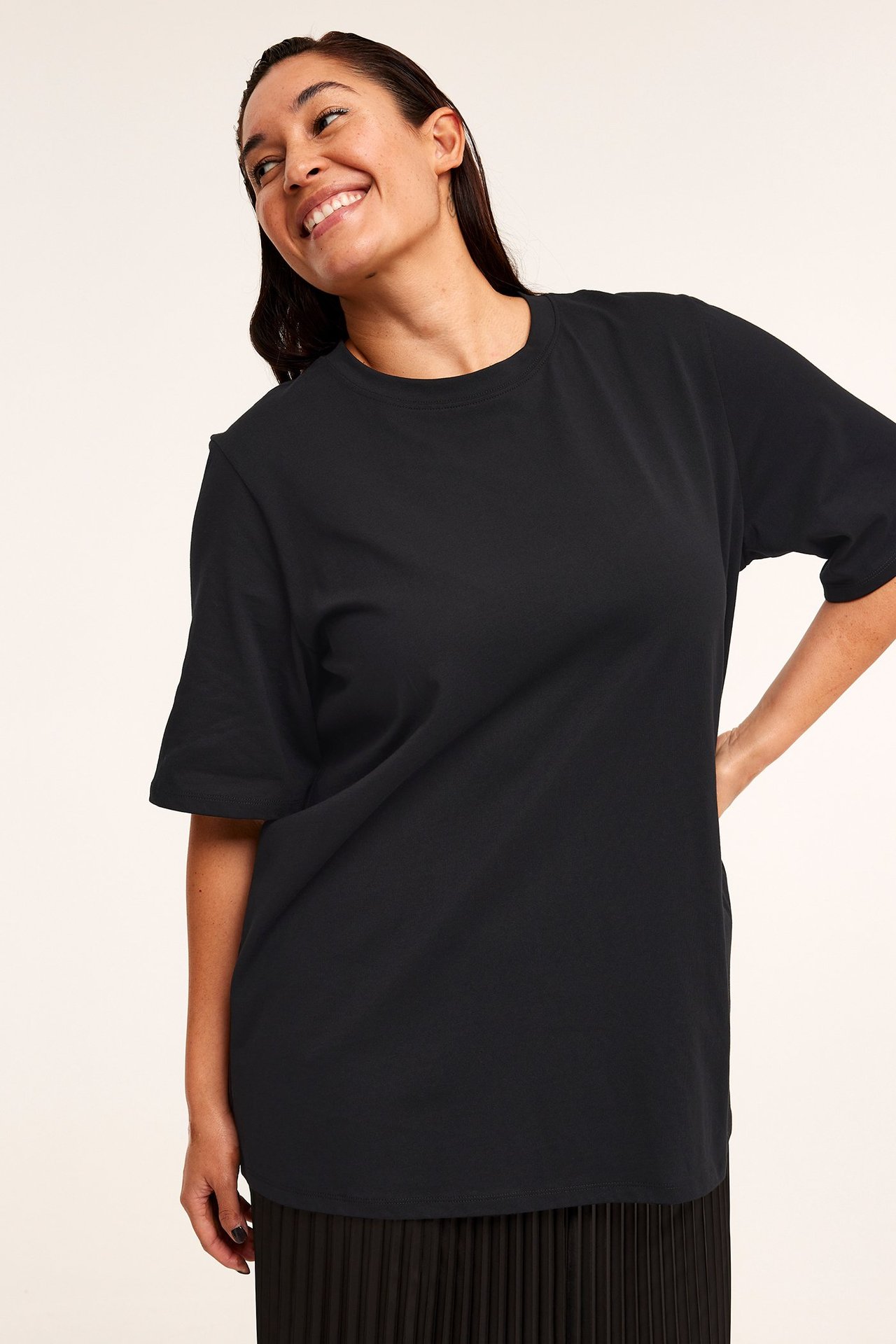 Edestäpäin hymyilevä nainen yllään musta rento puuvillainen t-paita ja musta laskostettu hame.