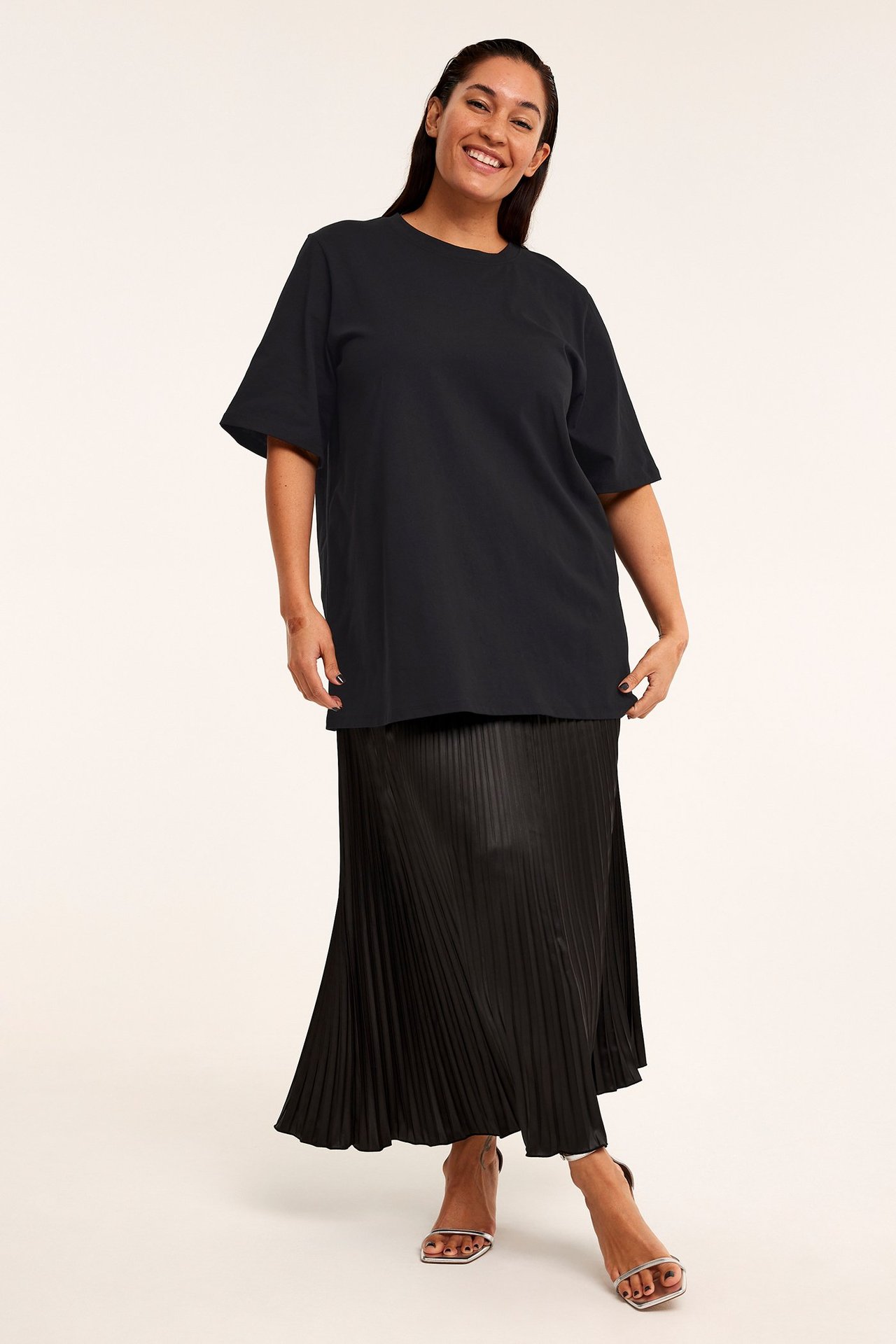 Widok z przodu: kobieta w czarnym, luźnym bawełnianym t-shircie, czarnej plisowanej spódnicy maxi i srebrnych sandałach.