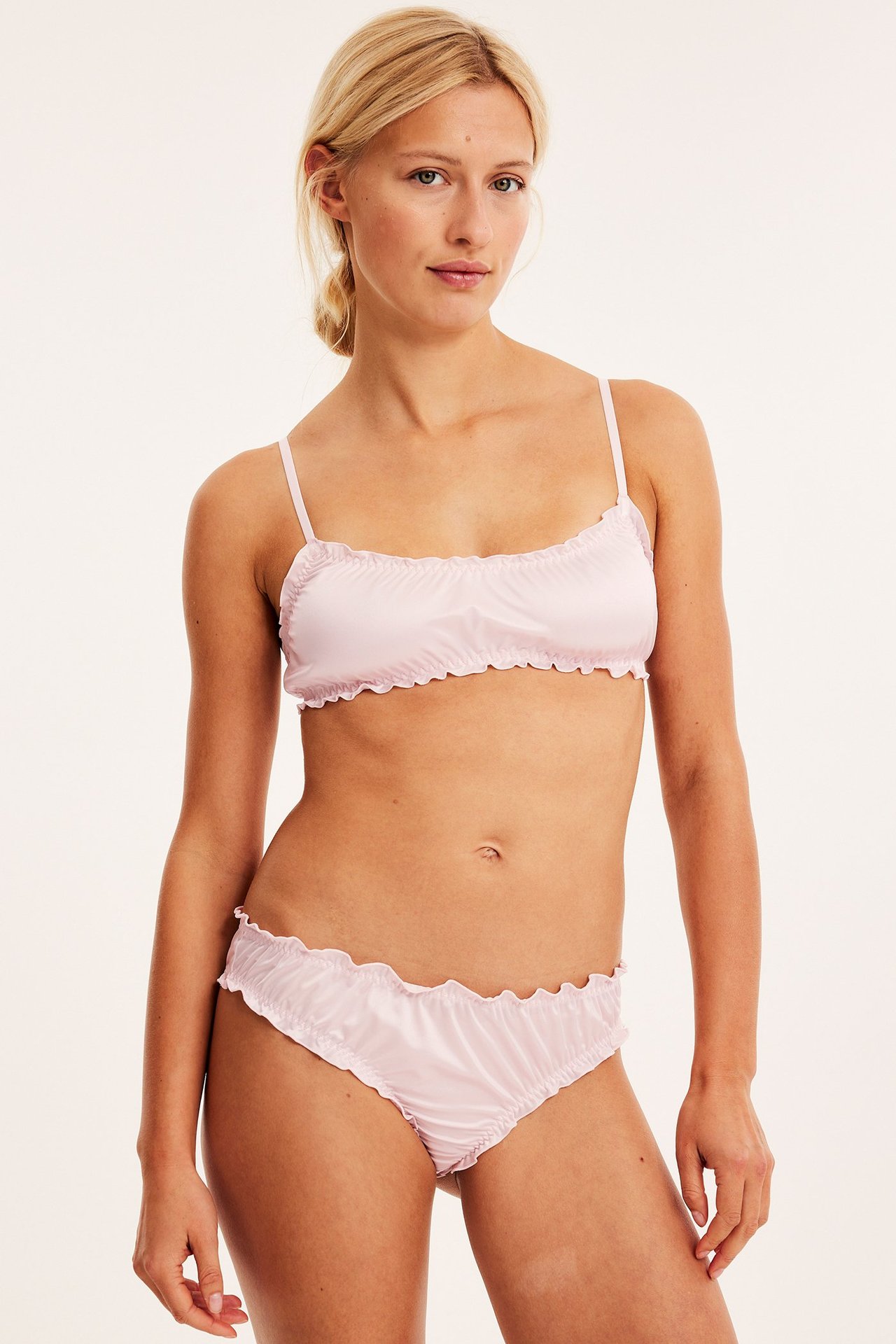 Framifrån, kvinna i ljusrosa satinbralette med volangkanter och matchande trosa.