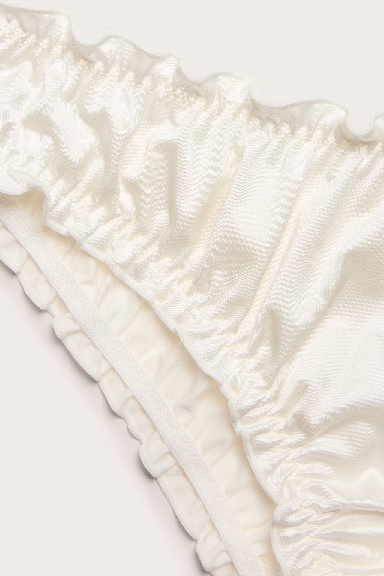 Nærbilde av en offwhite sateng brazilian-truse med volangkanter og rynkede detaljer.