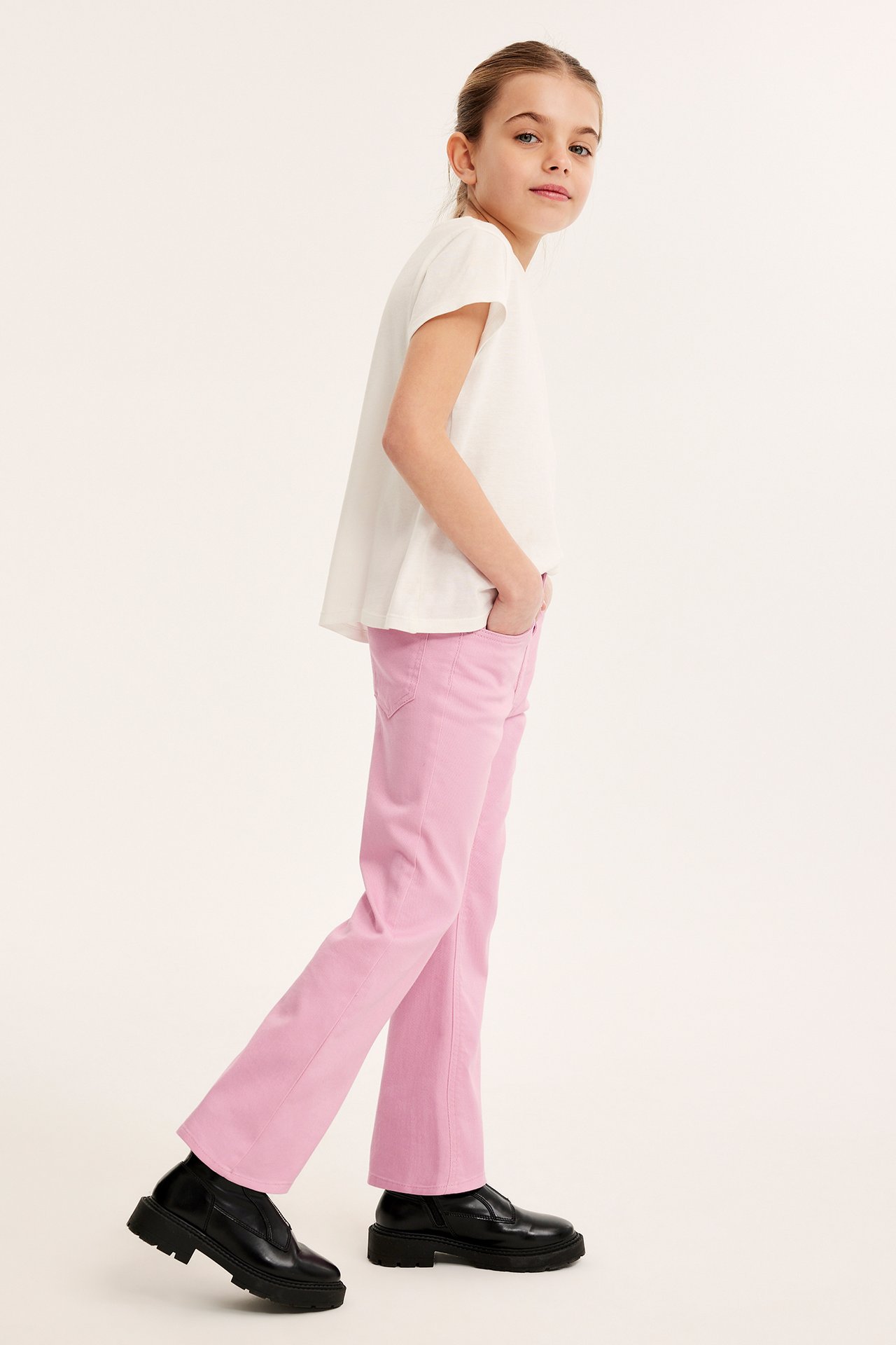 Sidovy av ett barn i rosa bootcut-jeans, en vit t-shirt och svarta grova skor.