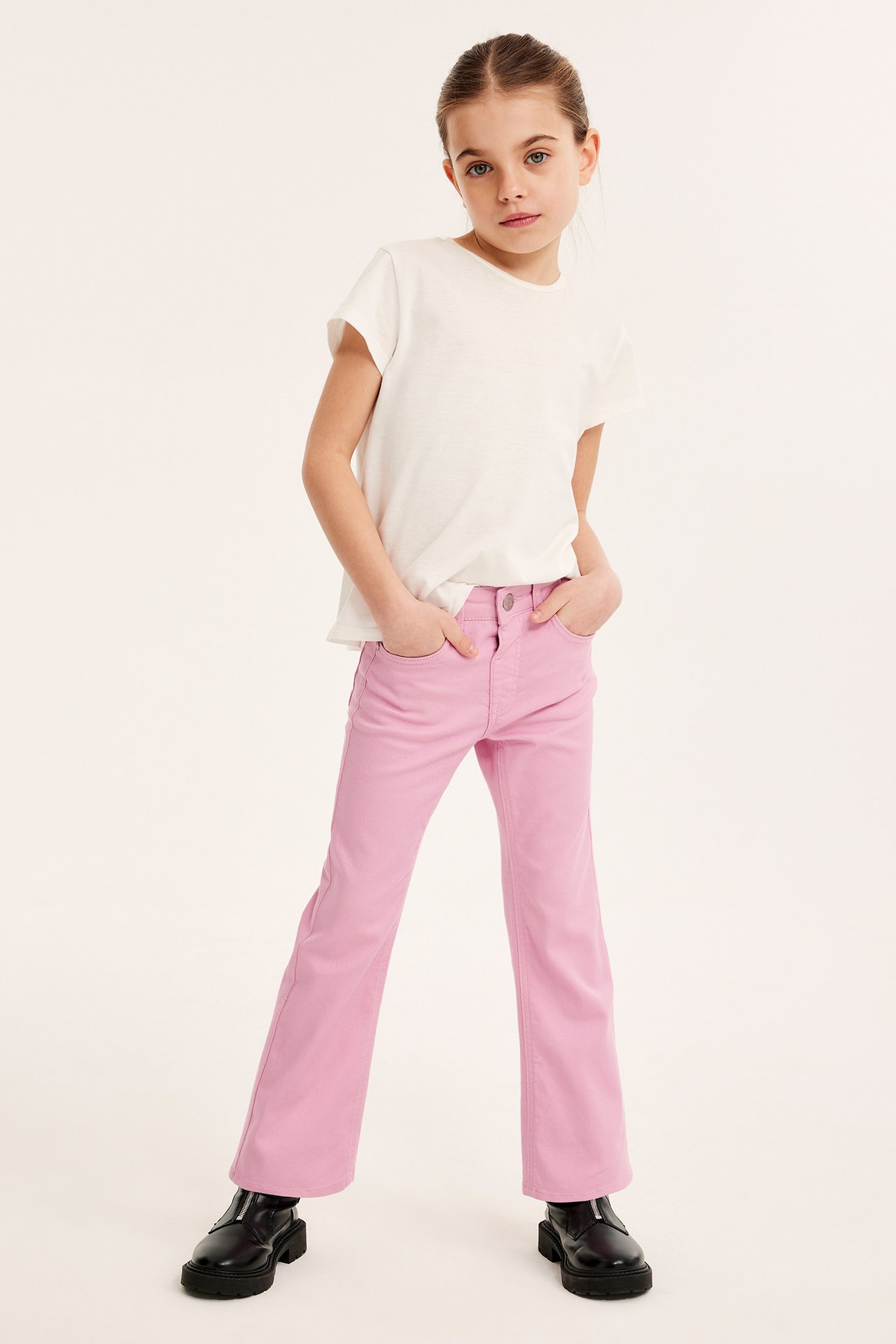 Framsida av ett barn i rosa bootcut-jeans med tryckknapp, en vit t-shirt och svarta kängor.