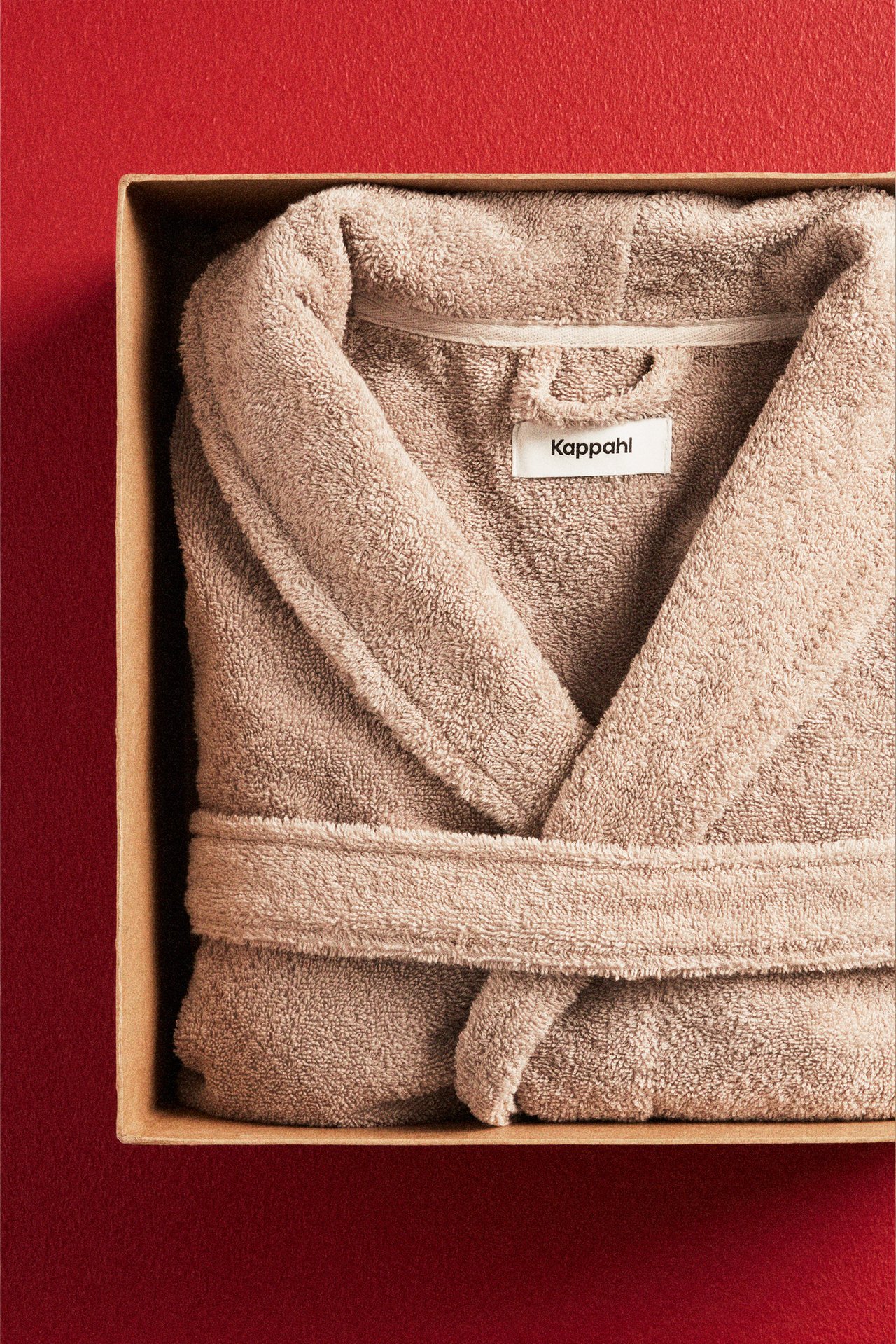 Närbild uppifrån på en vikt beige enfärgad bomullsfrottémorgonrock med skärp i en brun låda.