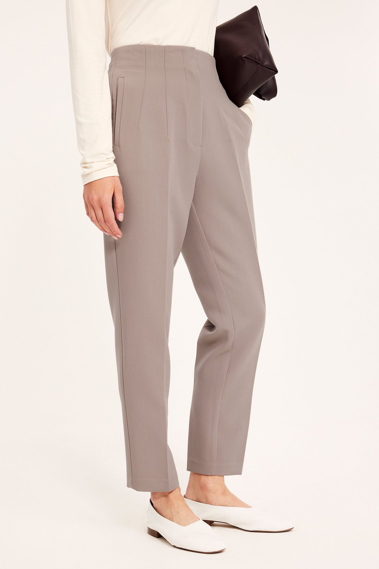 Kvinne i mørk beige smale høylivsbukser, off-white topp, hvite flate sko, holder brun clutch.