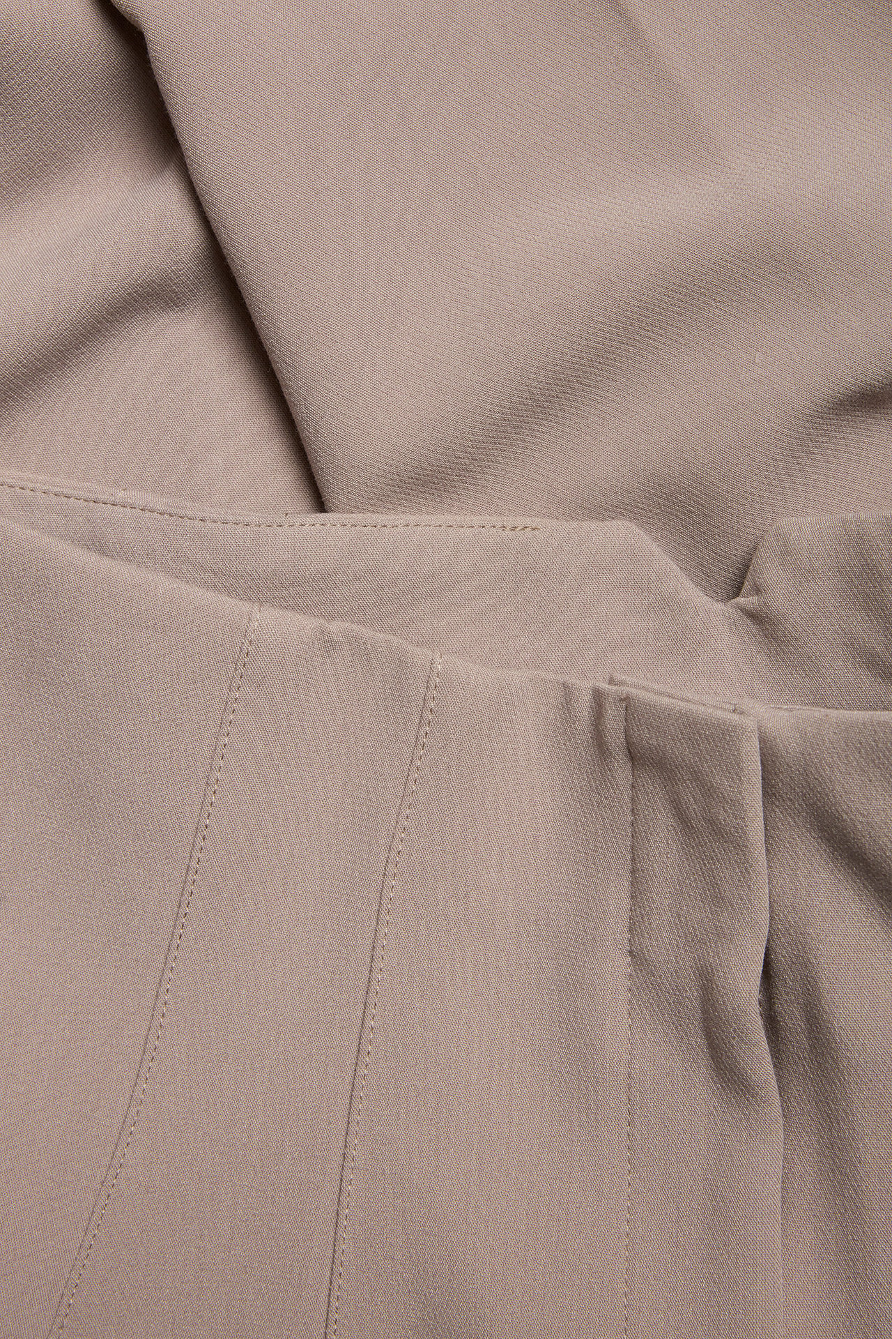 Zbliżenie na ciemnobeżowe spodnie garniturowe z wysokim stanem, zakładkami i widocznymi szwami.