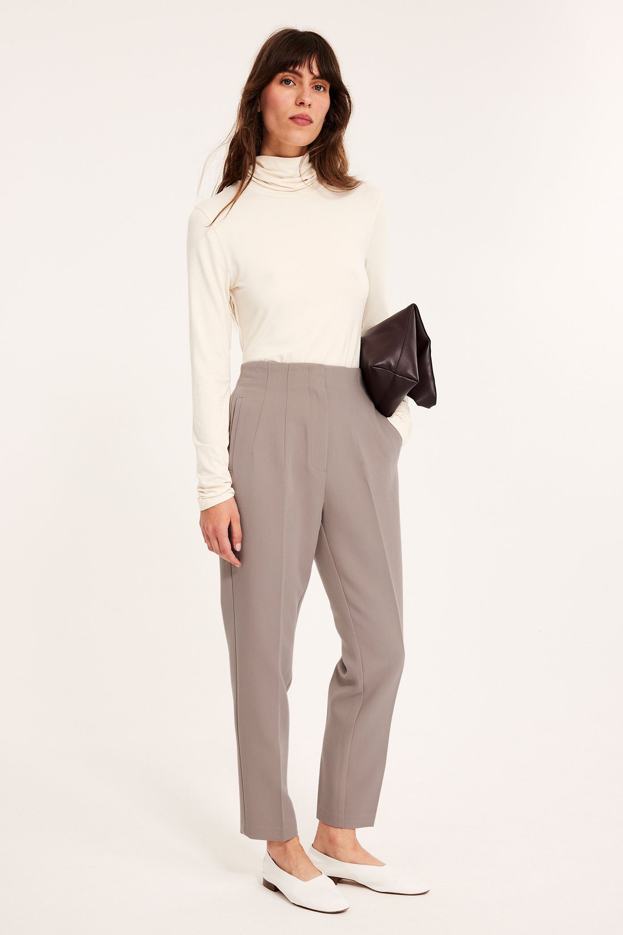 Widok z przodu: Kobieta w ciemnobeżowych, wysokich, dopasowanych spodniach garniturowych, kremowym golfie, ciemnobrązowej kopertówce, białych balerinach.