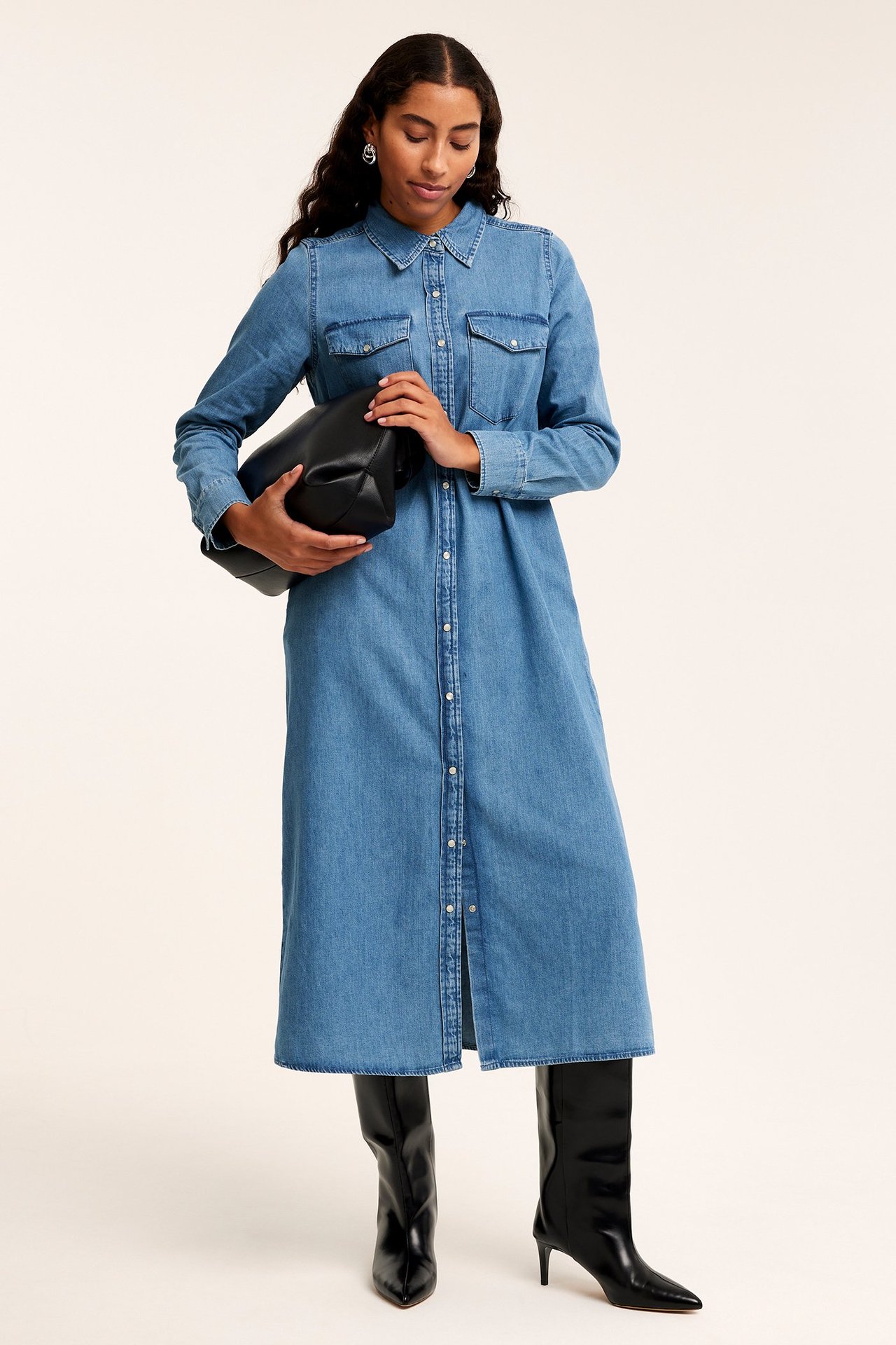 Framifrån: kvinna i lång blå jeansskjortklänning, svarta stövlar, håller svart clutchväska.