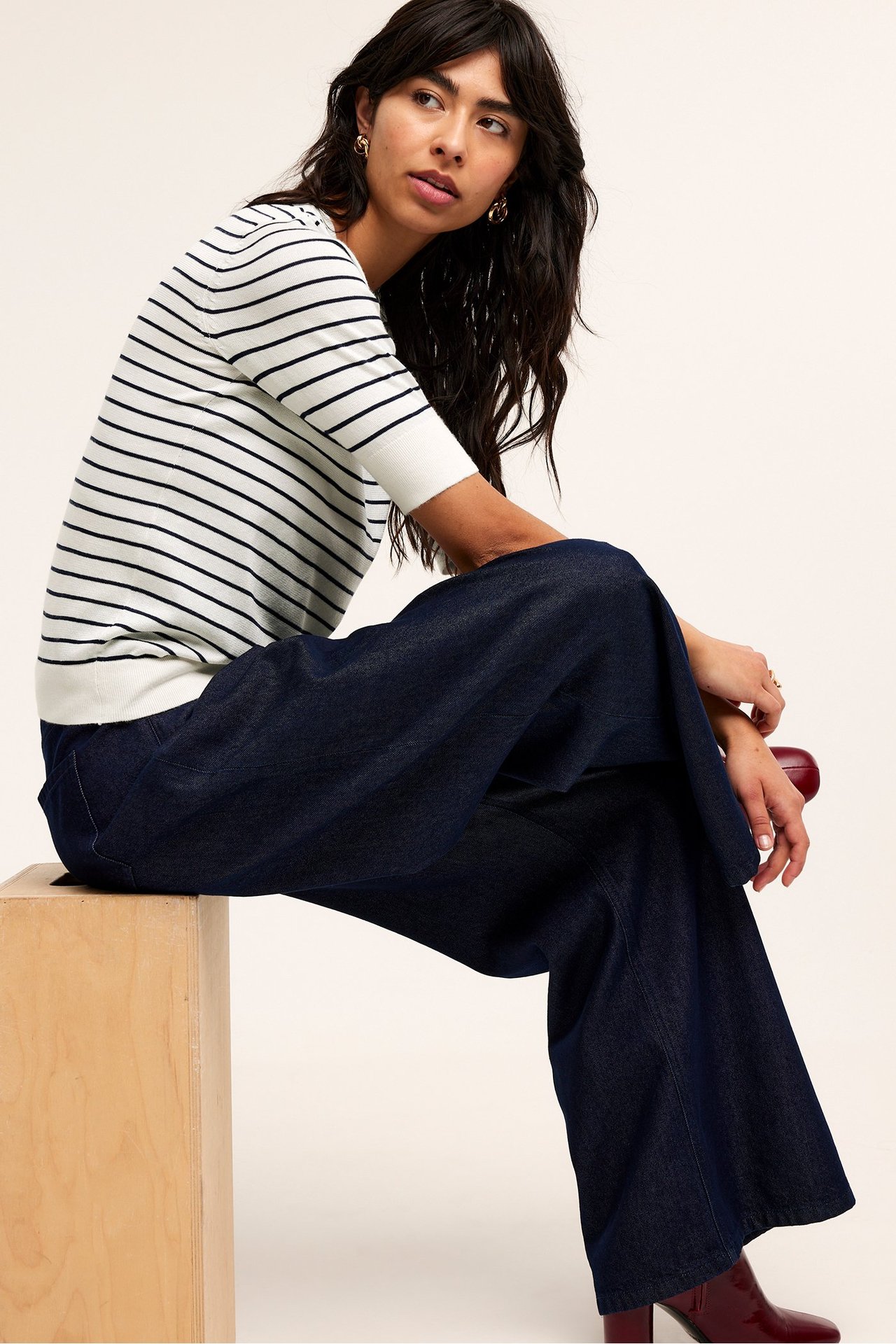 Edestä, nainen valkoisessa raidallisessa hienoneulepuserossa, tummansinisissä leveälahkeisissa housuissa ja tummanpunaisissa saappaissa.