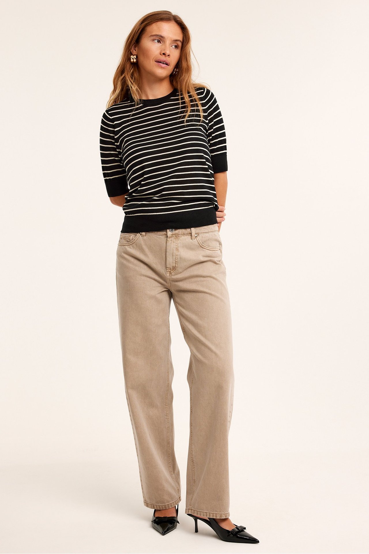 Edestä: nainen mustavalkoraidallisessa lyhythihaisessa neuleessa, beigenvärisissä leveissä housuissa ja mustissa koroissa.