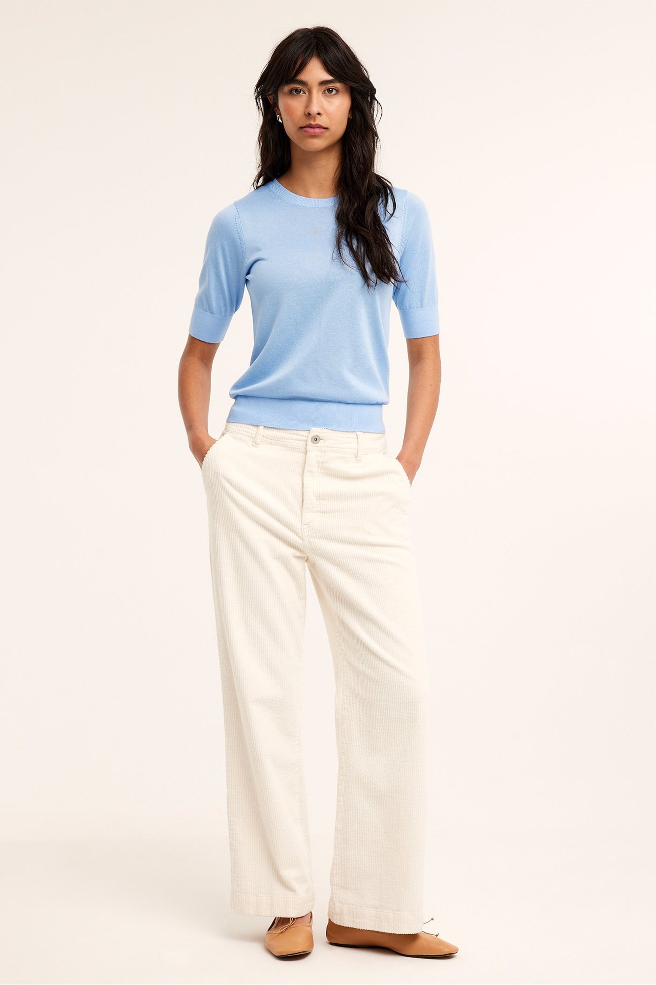 Front view: nainen yllään vaaleansininen lyhythihainen neulepusero, kermanväriset leveälahkeiset vakosamettihousut, ruskeat ballerinat.