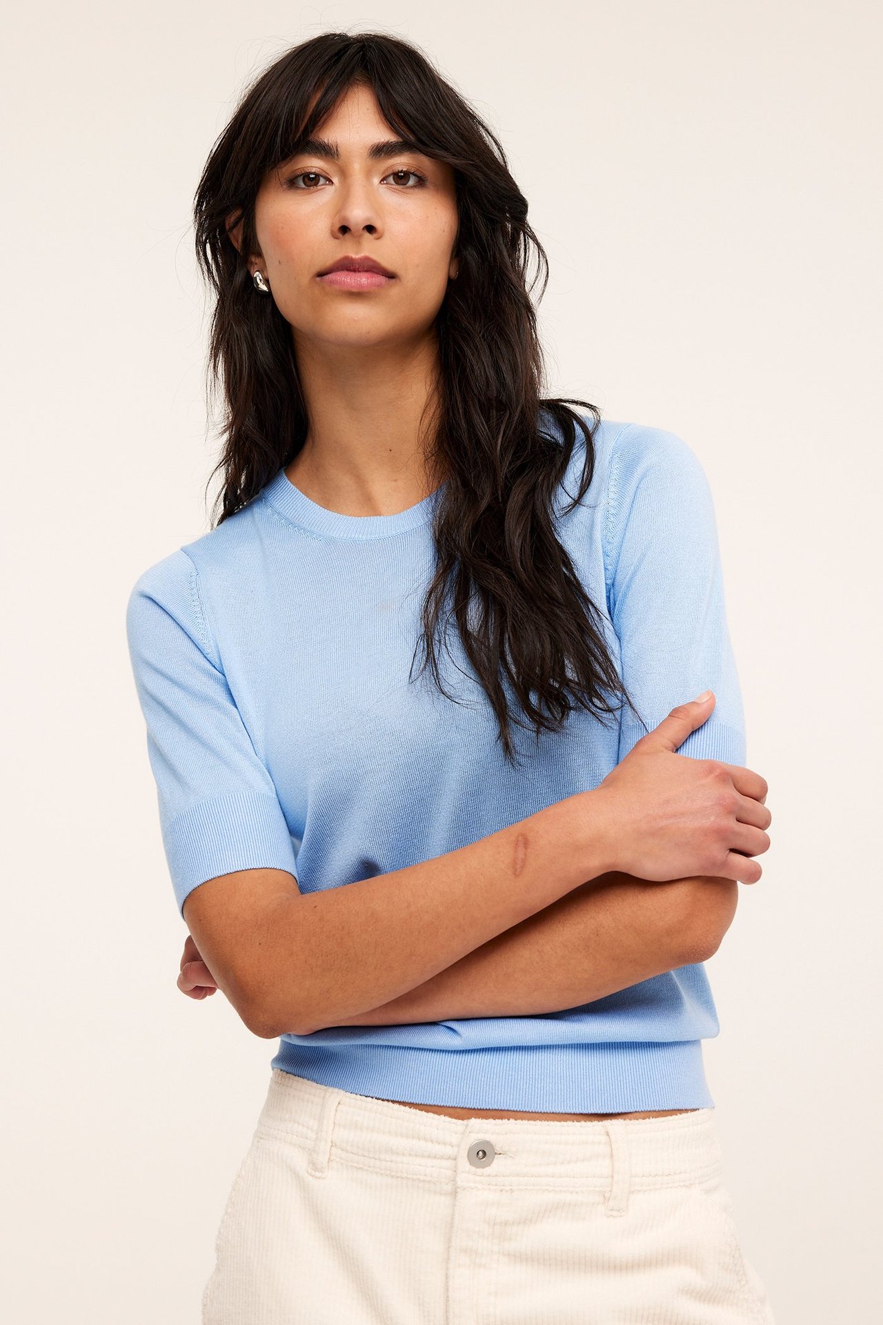 Edestä kuvattu nainen vaaleansinisessä lyhythihaisessa neuleessa ja kermanvärisissä vakosamettihousuissa.