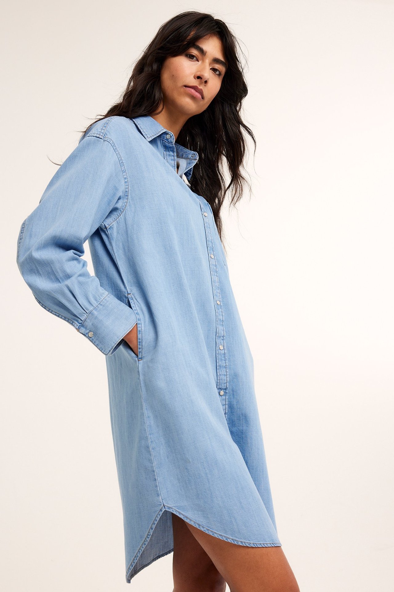 Edestä: Nainen yllään väljä vaaleansininen farkkupaitamekko pitkillä hihoilla ja sivutaskuilla.