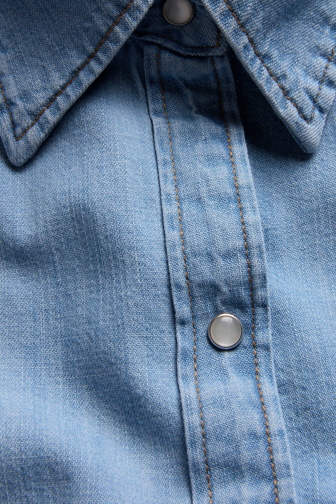 Zbliżenie: Jasnoniebieska dżinsowa sukienka koszulowa z kołnierzem, listwą guzikową, srebrnymi zatrzaskami i brązowymi szwami.