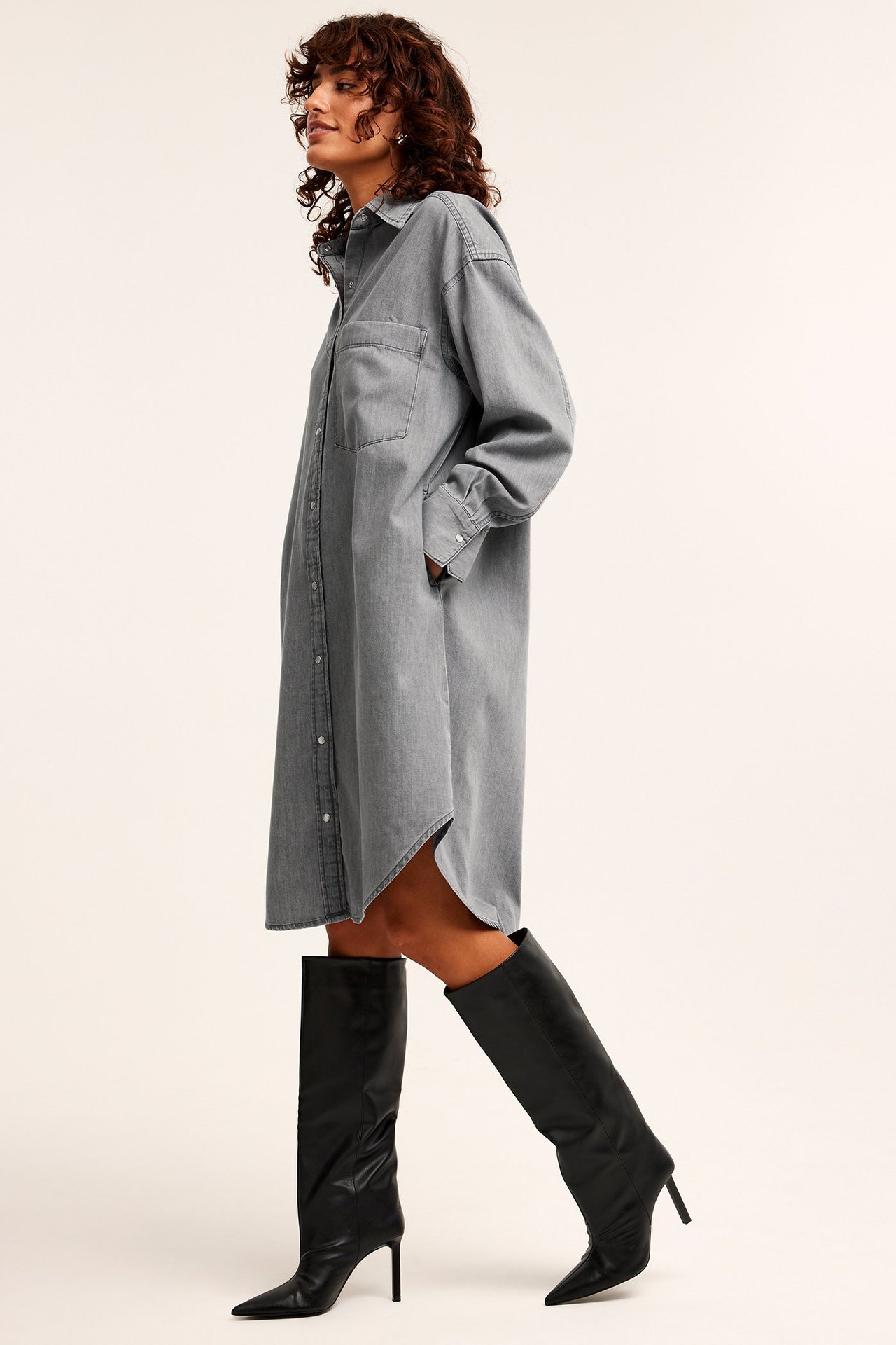 Sidovy av kvinna i lång grå jeansskjortklänning med knappar och svarta knähöga stövlar.