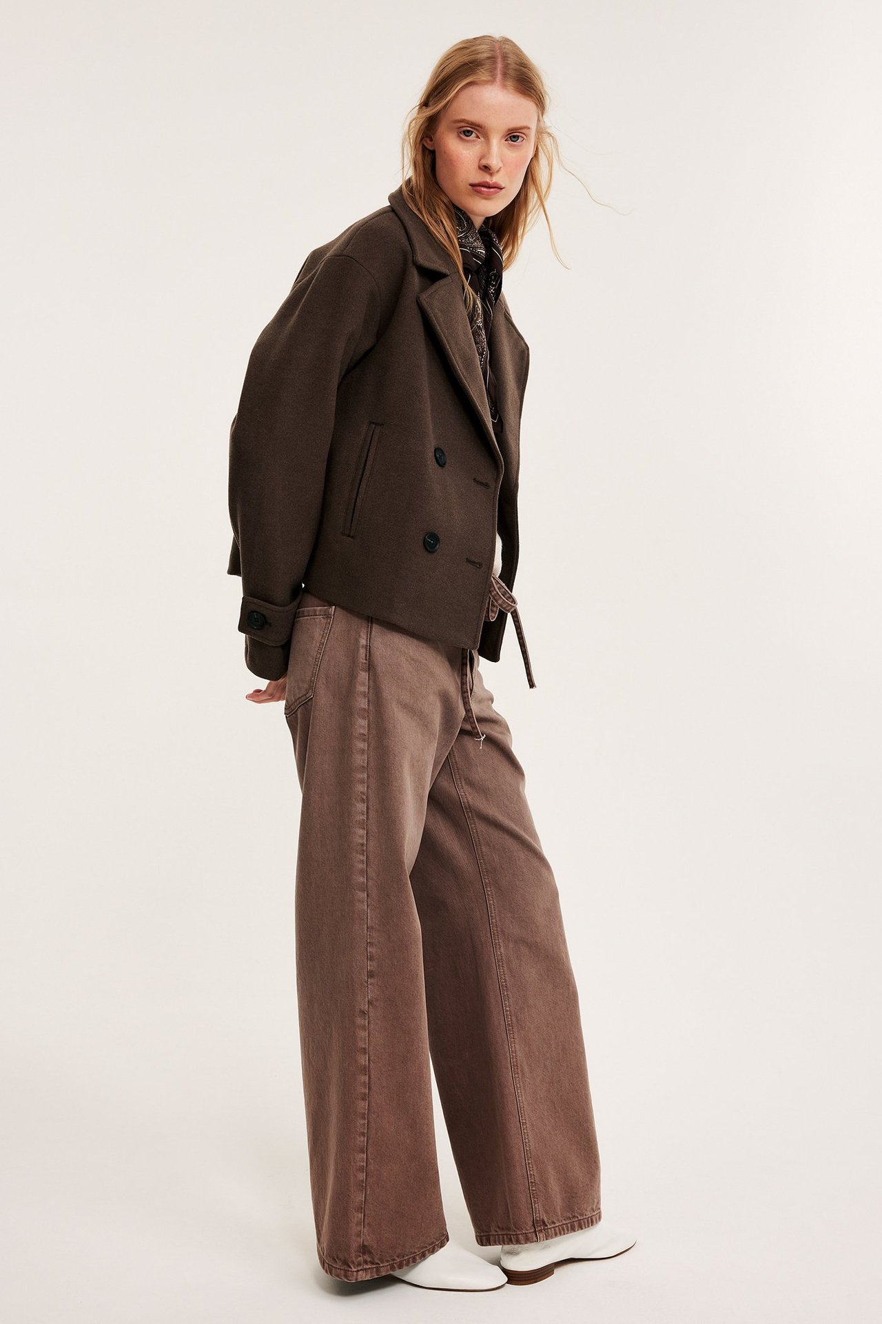 Forfra: kvinne i mørkebrun kort peacoat, brune vide bukser, mønstret skjerf, hvite sko.