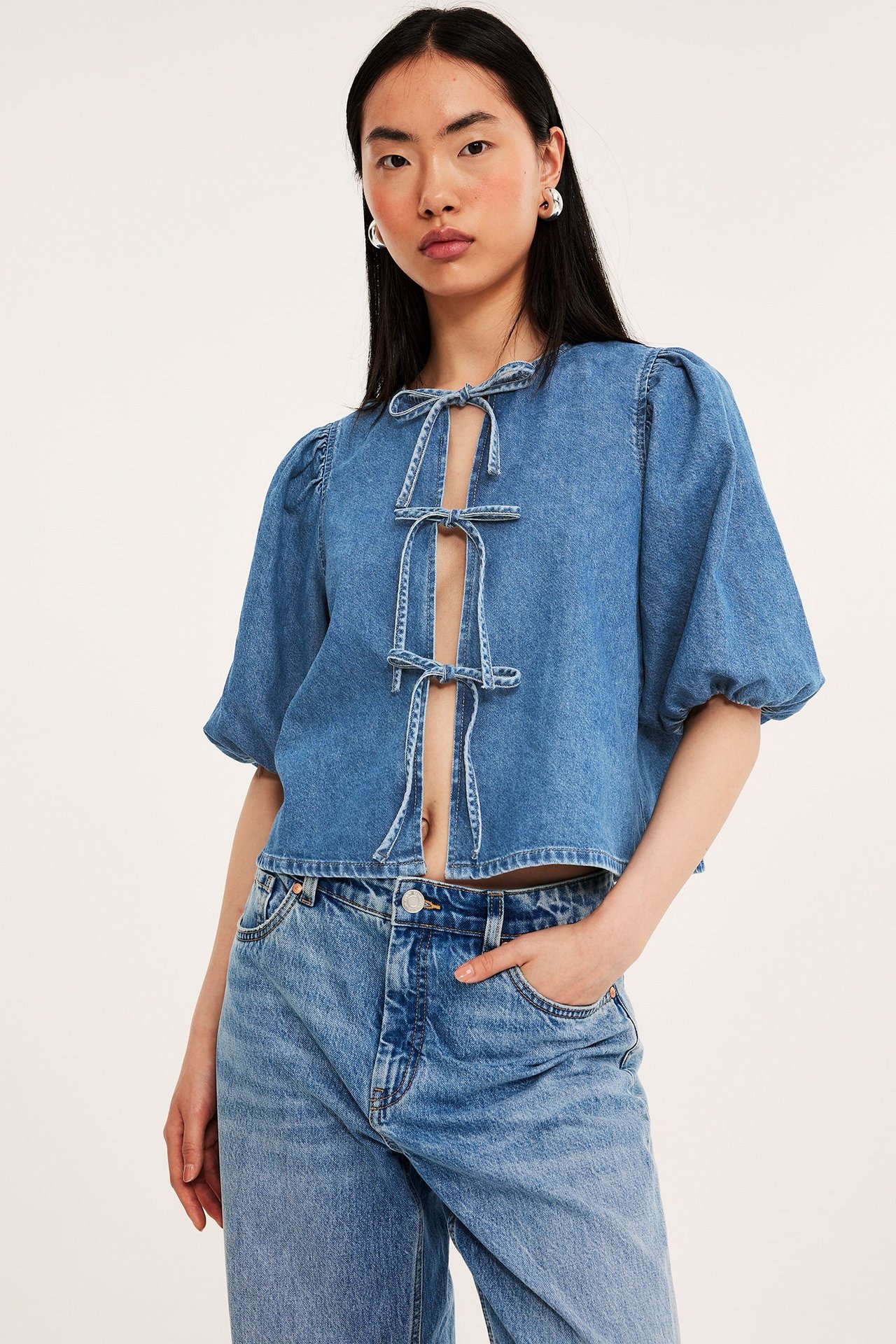 Front: Kvinne i blå denimbluse med puffermer og knyting foran, lyseblå jeans og sølvfargede ringøreringer.