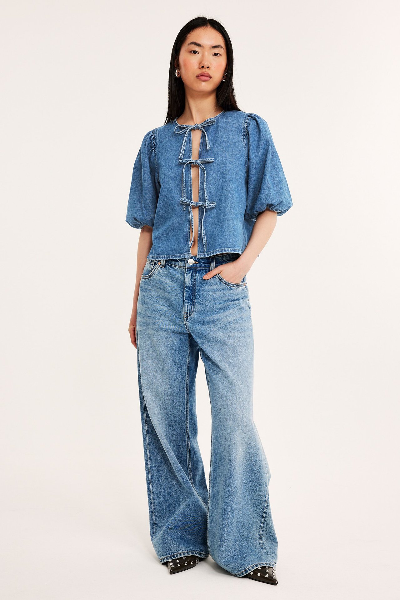 Widok z przodu: Kobieta w jasnoniebieskiej bluzce dżinsowej z bufiastymi rękawami, wiązaniami i szerokimi dżinsami.