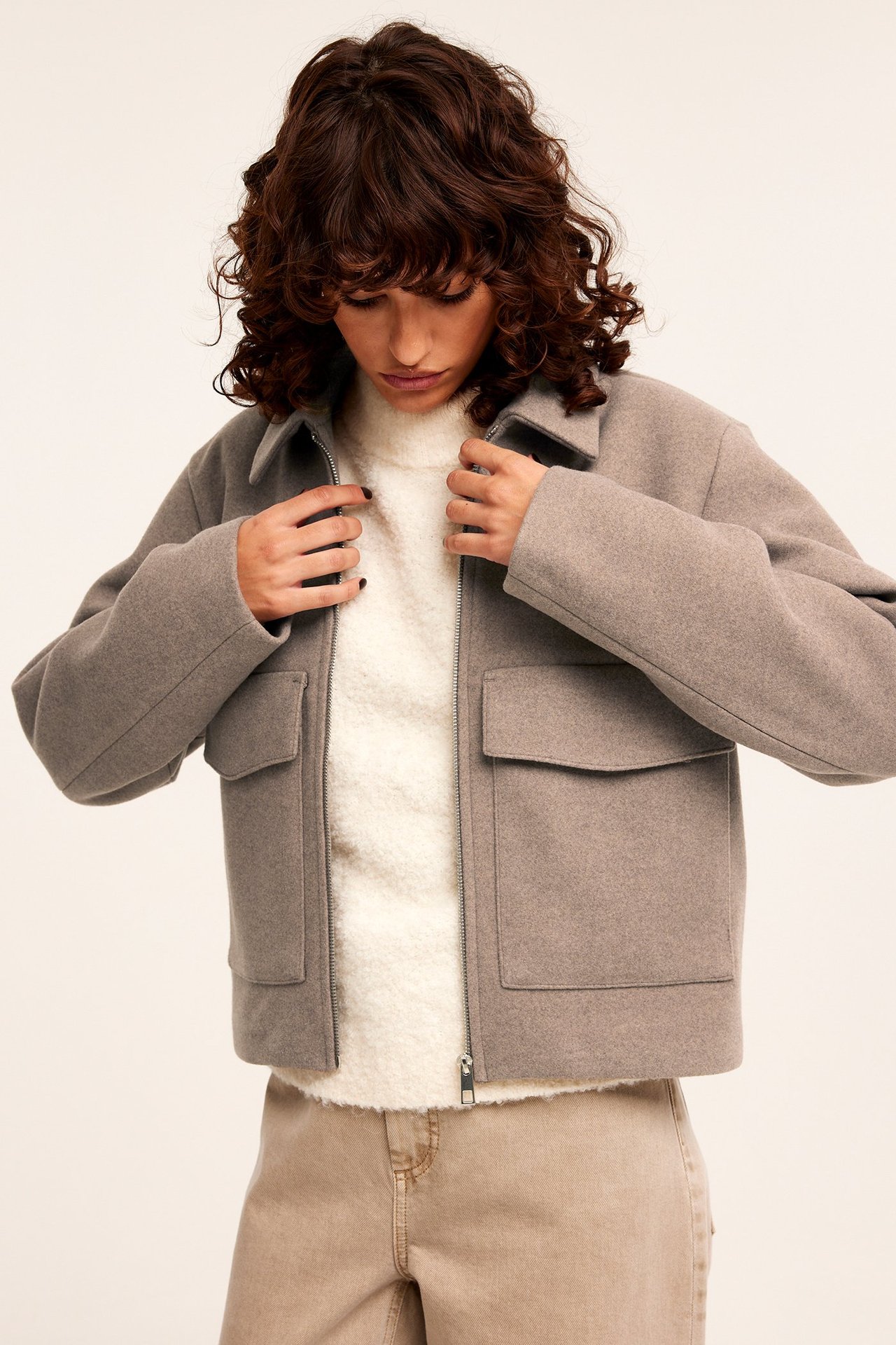 Edestä: Nainen beigessä meleeratussa huopatakissa vetoketjulla ja läppätaskuilla, valkoisen neuleen päällä.