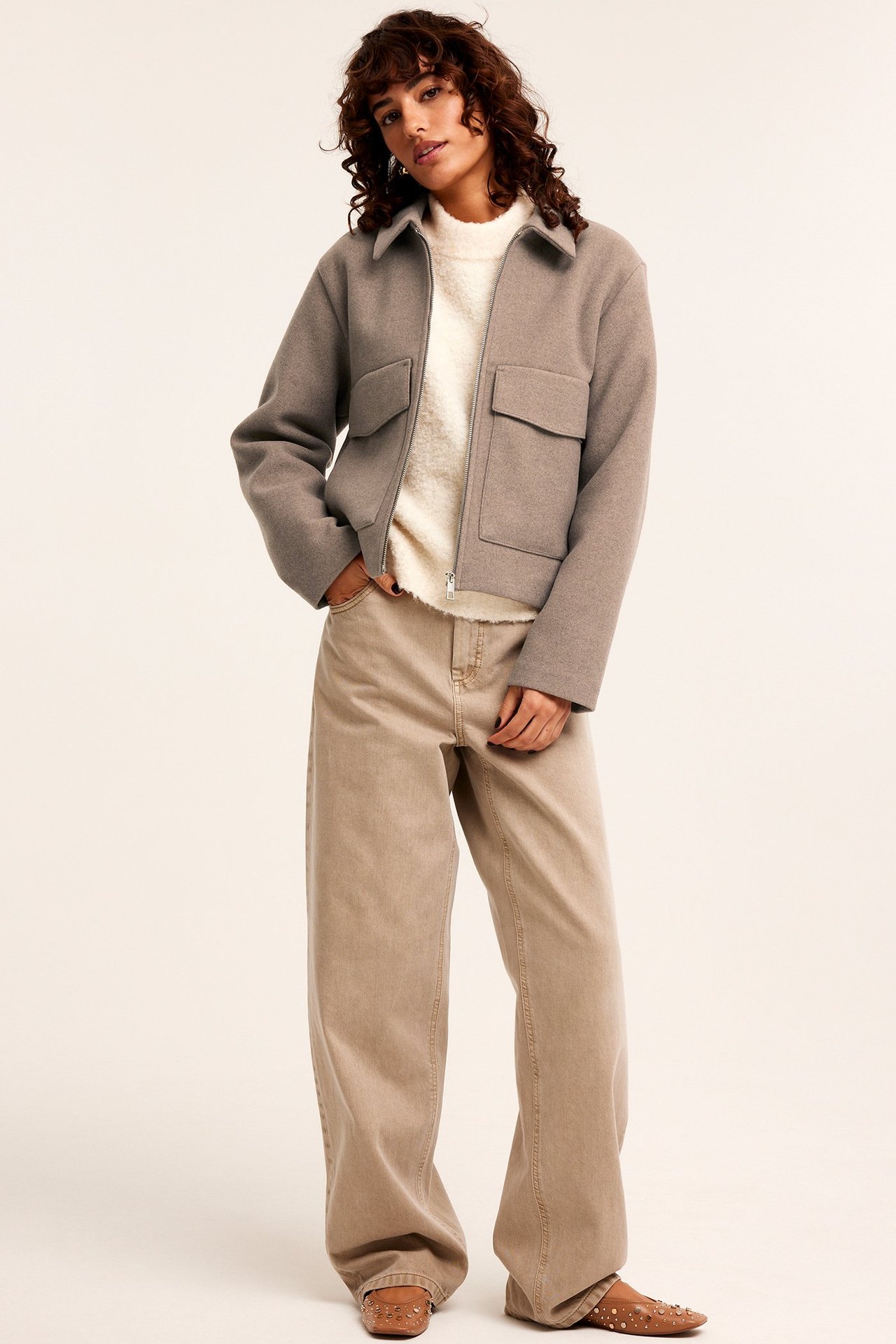 Widok z przodu: Kobieta w beżowej filcowej kurtce, białym swetrze, beżowych szerokich spodniach i brązowych nitowanych butach.